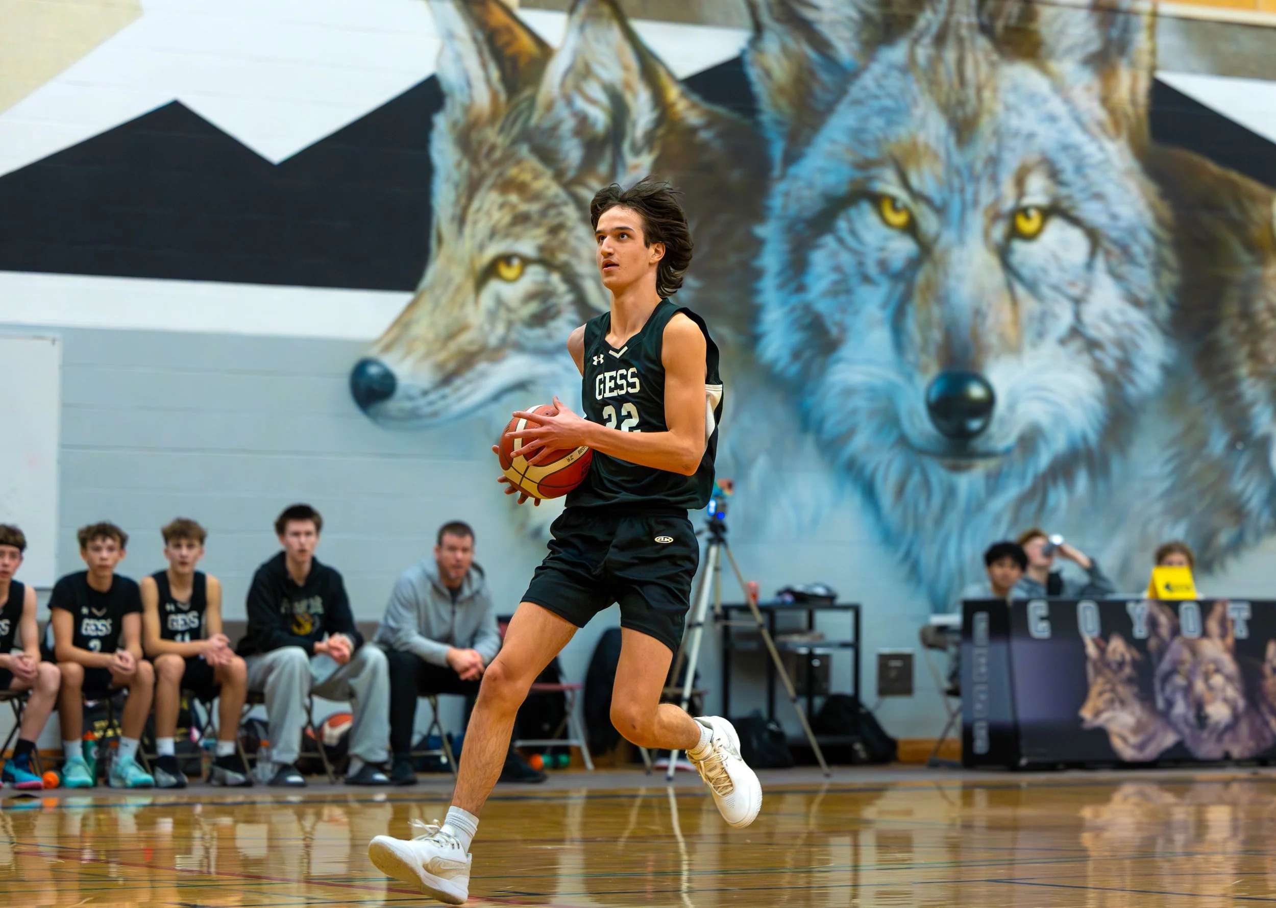 Young male basketball player in black uniform dribbling a basketball on the court with a wolf mural in the background and a seated team watching.