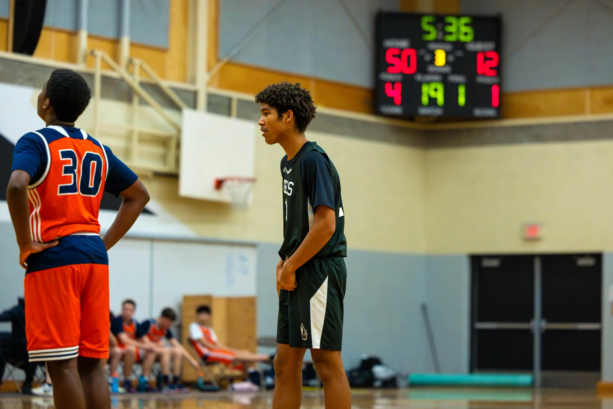 Two young basketball players on the court with a scoreboard showing scores and game time, one in a black and green uniform and the other in a red and blue uniform, with teammates seated on the bench in the background.