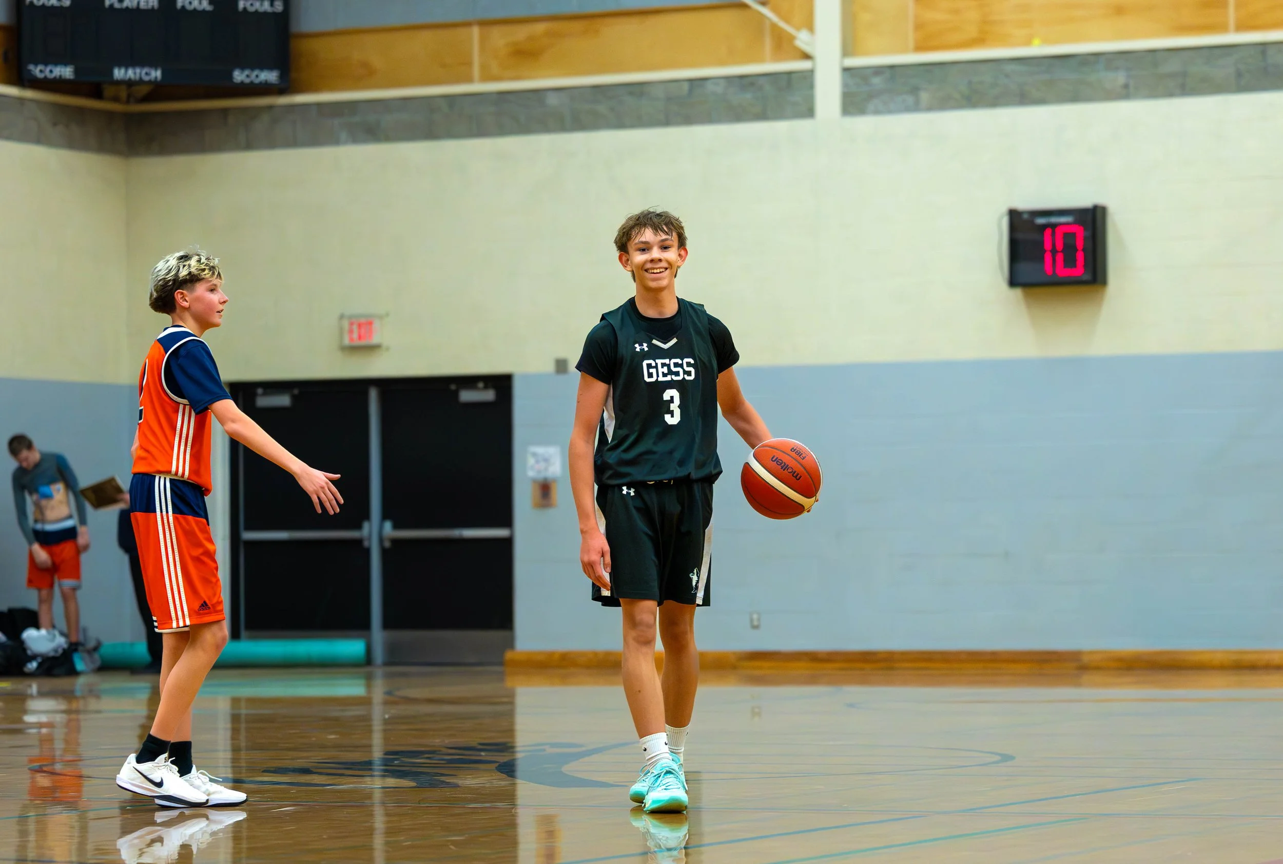 A young boy in a black GESS basketball uniform holding a basketball on an indoor court, with another boy in a blue and orange uniform in the background.