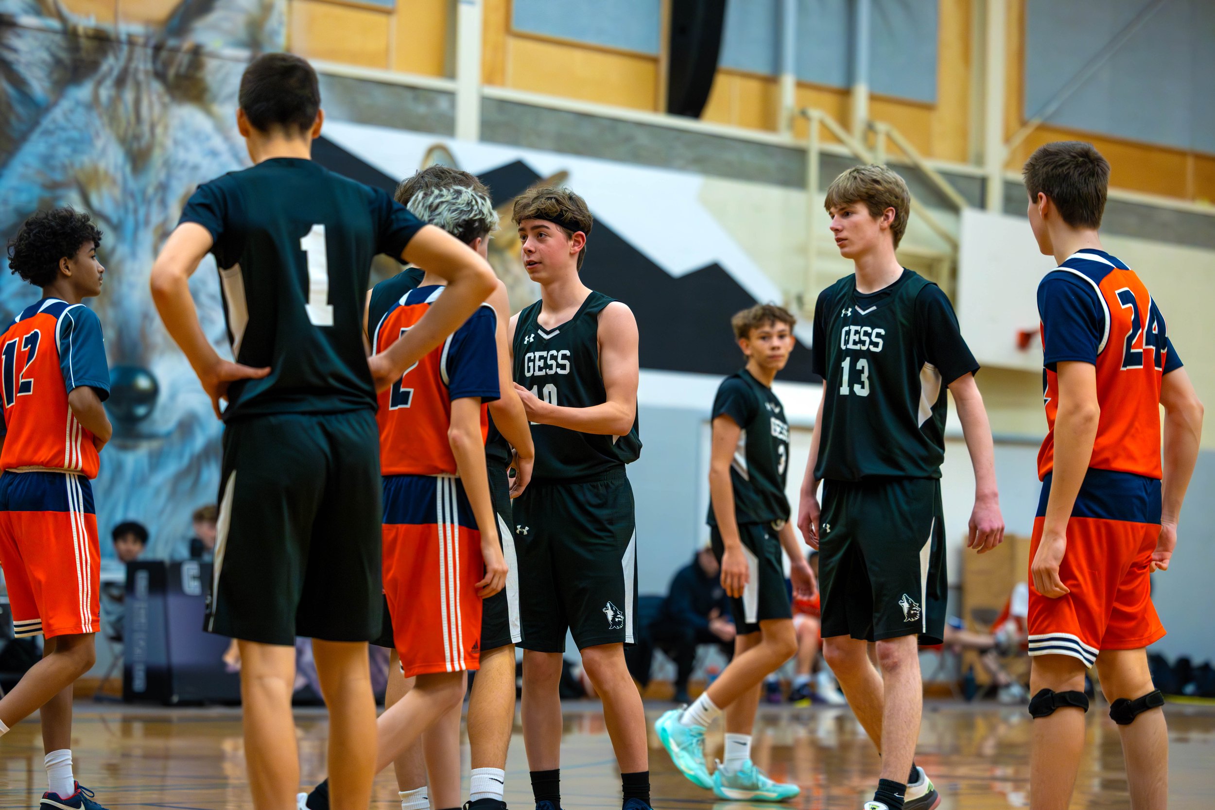 Youth basketball game with players in black and orange jerseys, on an indoor court, some players talking while others walk or prepare for the game.