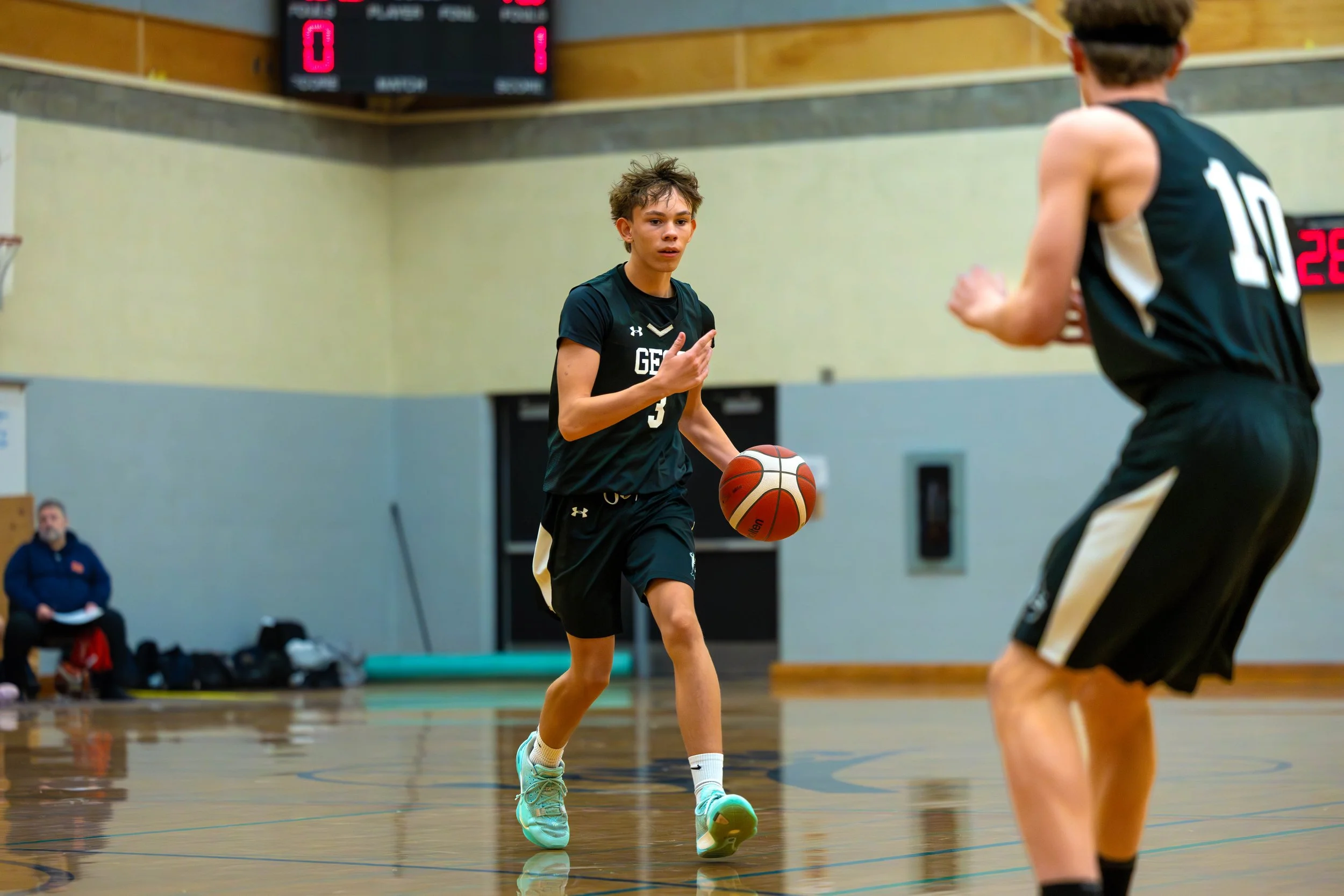 Two young basketball players in black uniforms practicing indoors, with one dribbling and the other preparing to pass, while a person sits on a bench in the background.