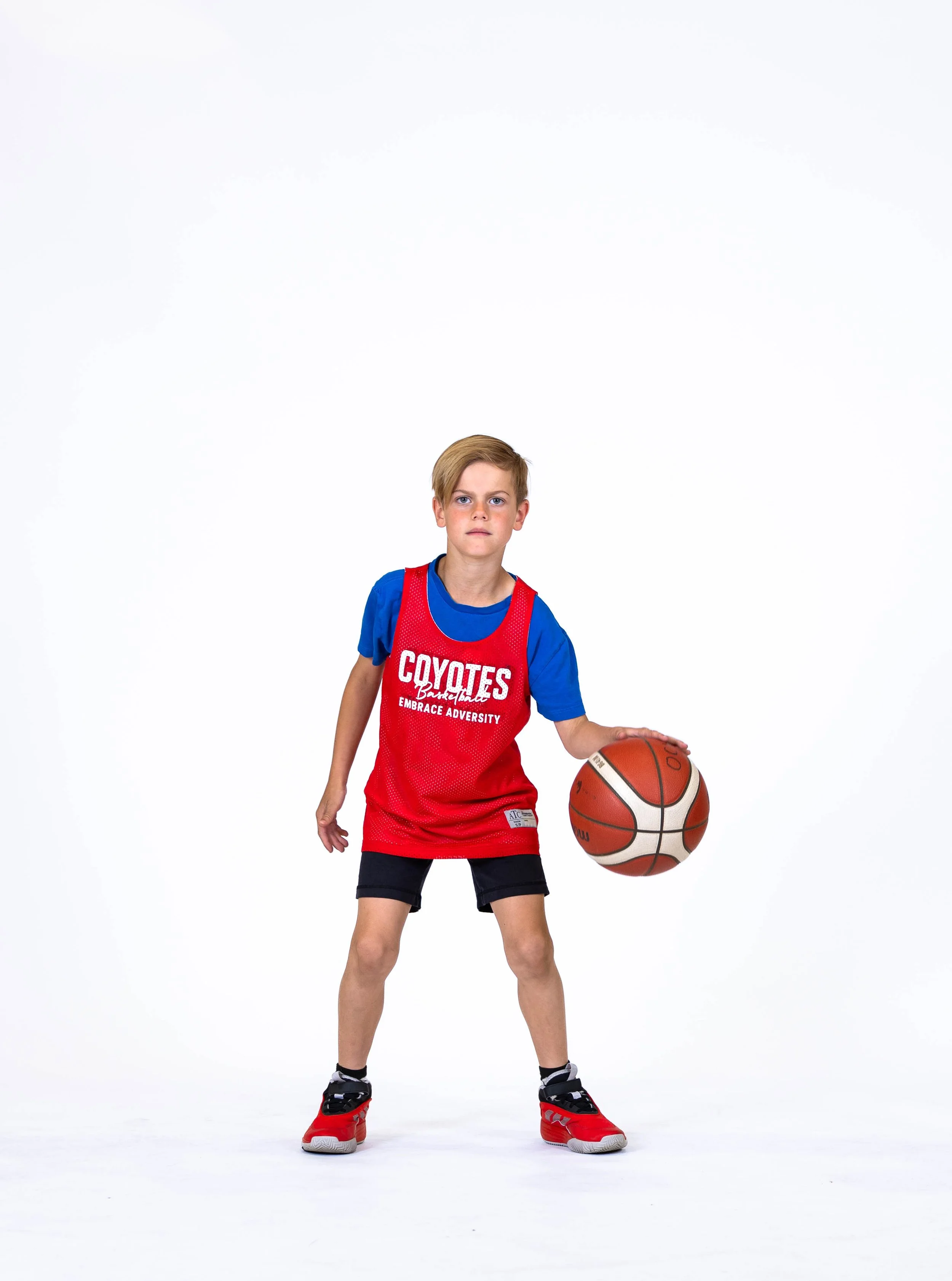 Young boy in a blue shirt with a red basketball jersey and black shorts standing with one hand on his hip and holding a basketball with his other hand against a white background.