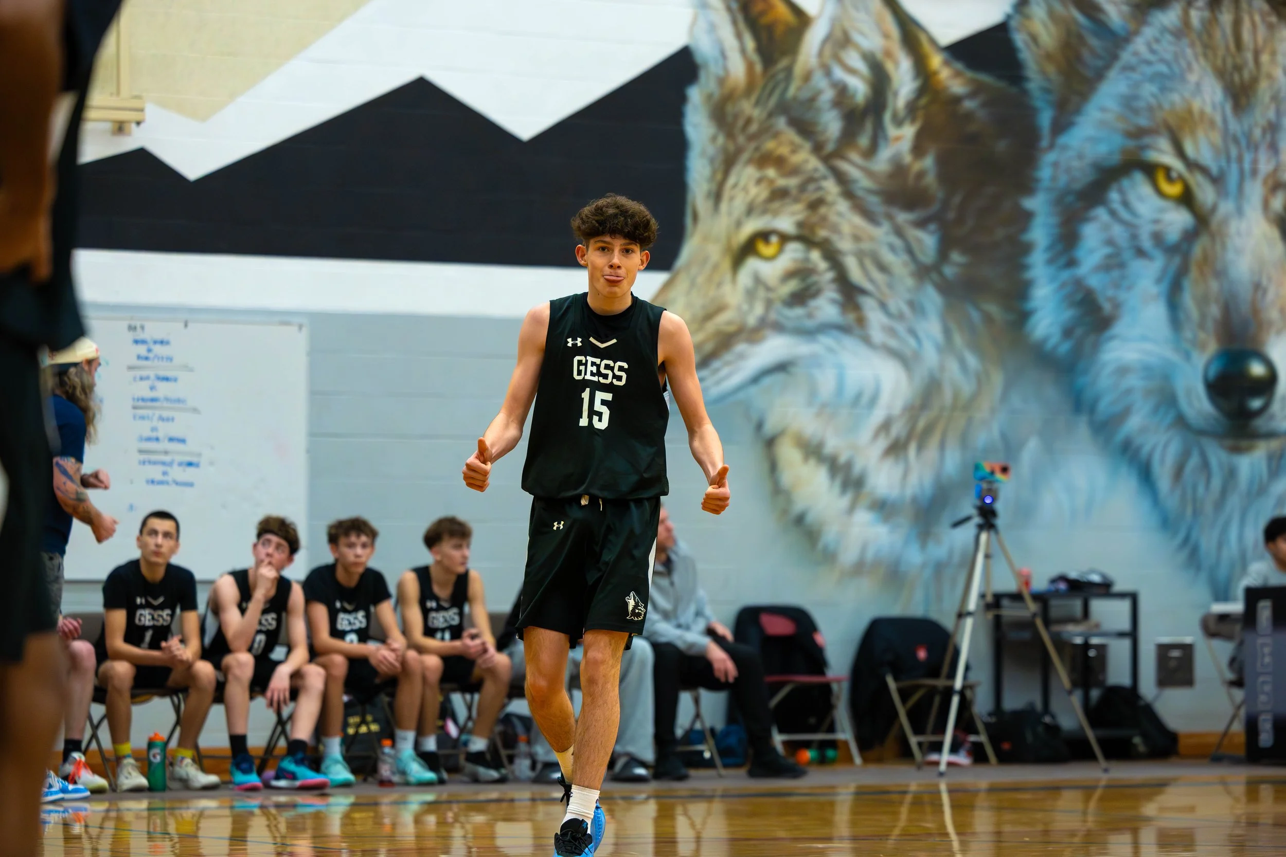A young male basketball player in a black GESS jersey with the number 15, standing on a gymnasium court with a large mural of wolves on the wall behind him. Several teammates sit on the bench watching, while a coach and a woman with tattoos and a hat