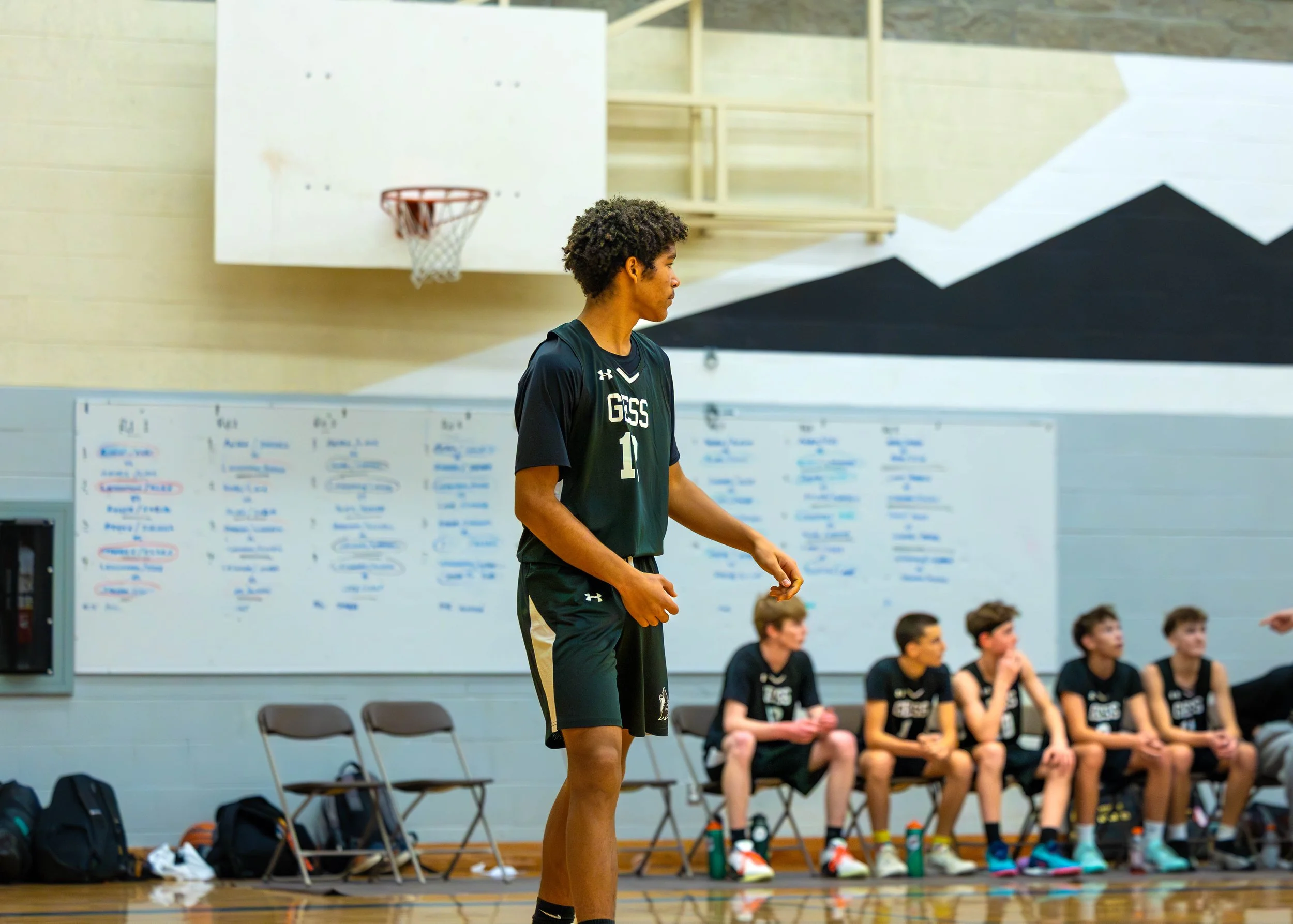 A young male basketball player standing on the court during a game or practice in a gymnasium, with other players sitting on the bench behind him and a whiteboard with notes in the background.