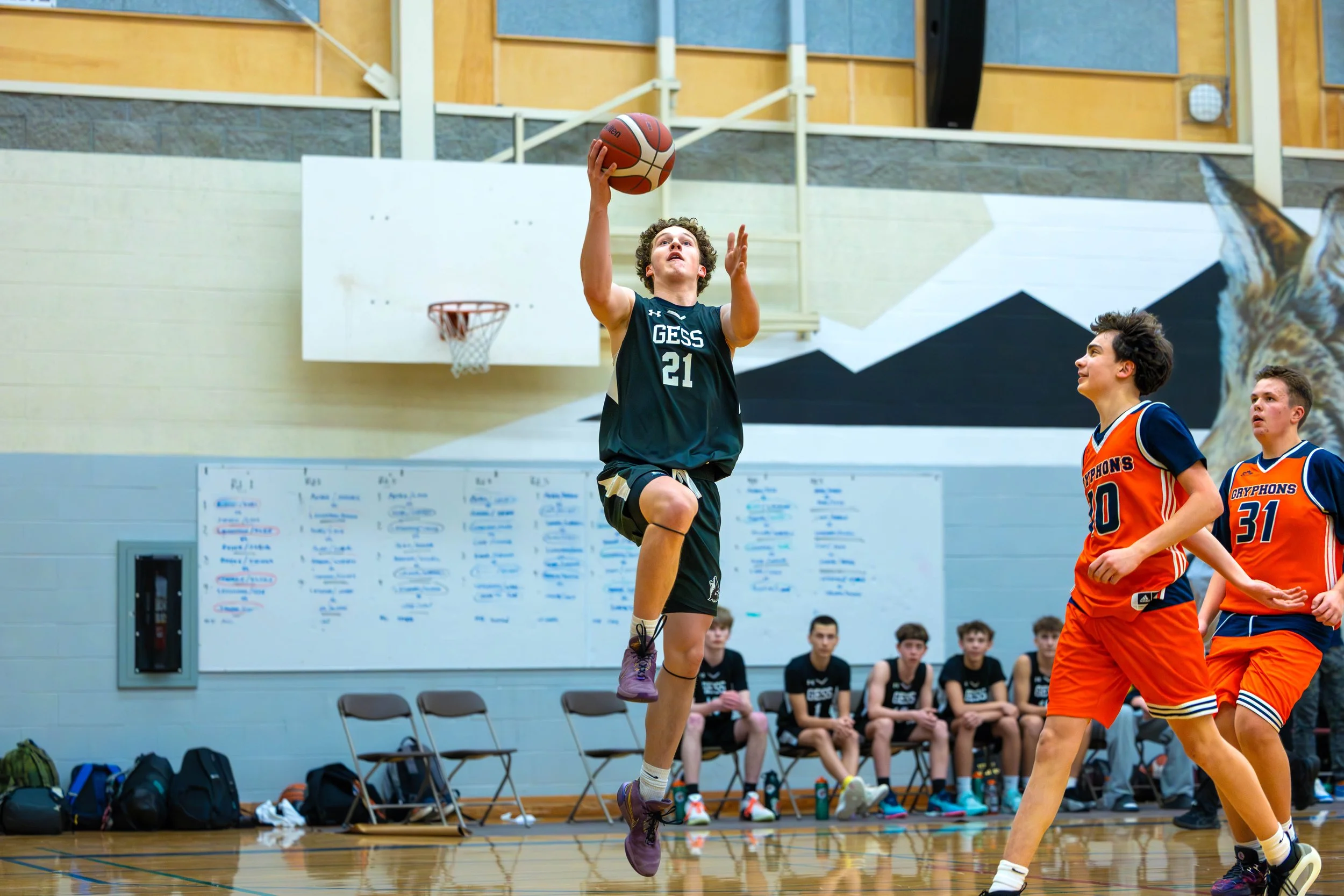 A basketball player in a green uniform jumps to make a shot near the basket while three players in orange uniforms defend during a game in a gymnasium.