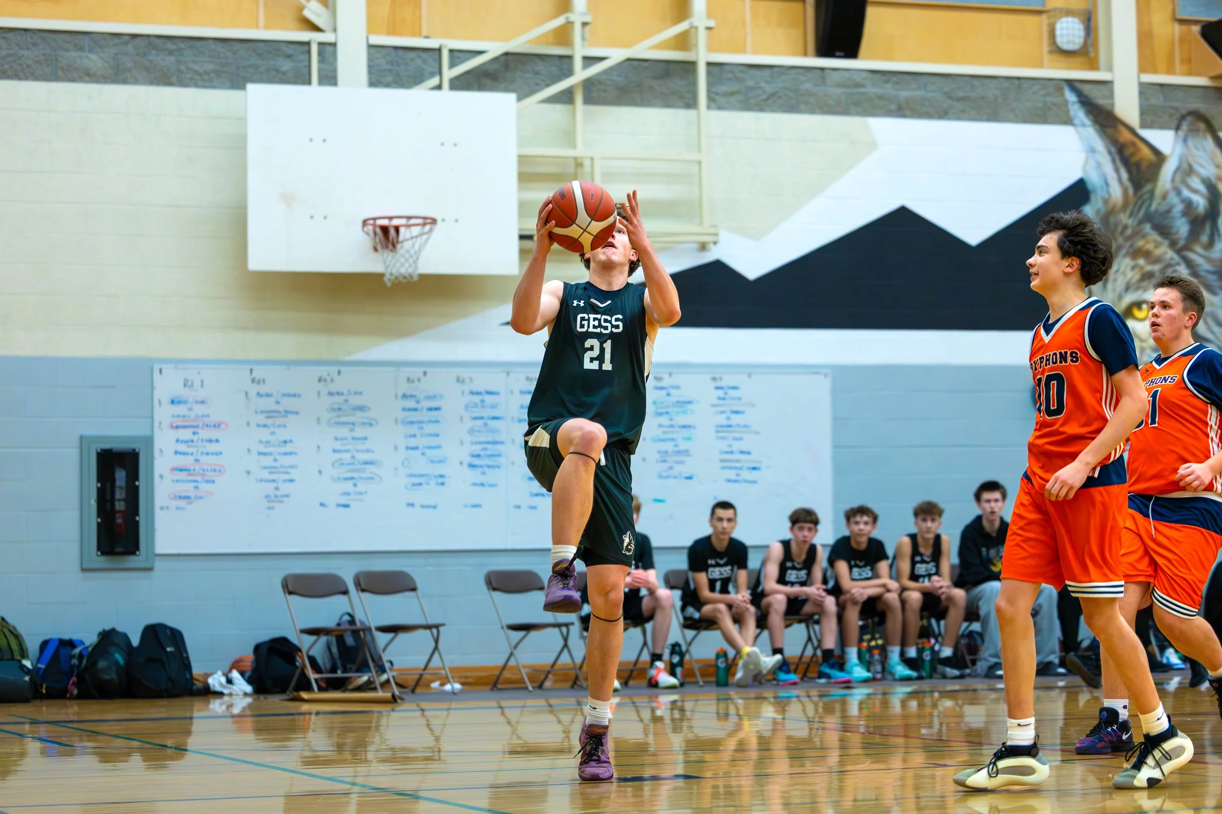 A young basketball player jumps with the ball, preparing to shoot inside a gym. Two other players wearing orange jerseys stand nearby, and several players sit on the bench in the background.