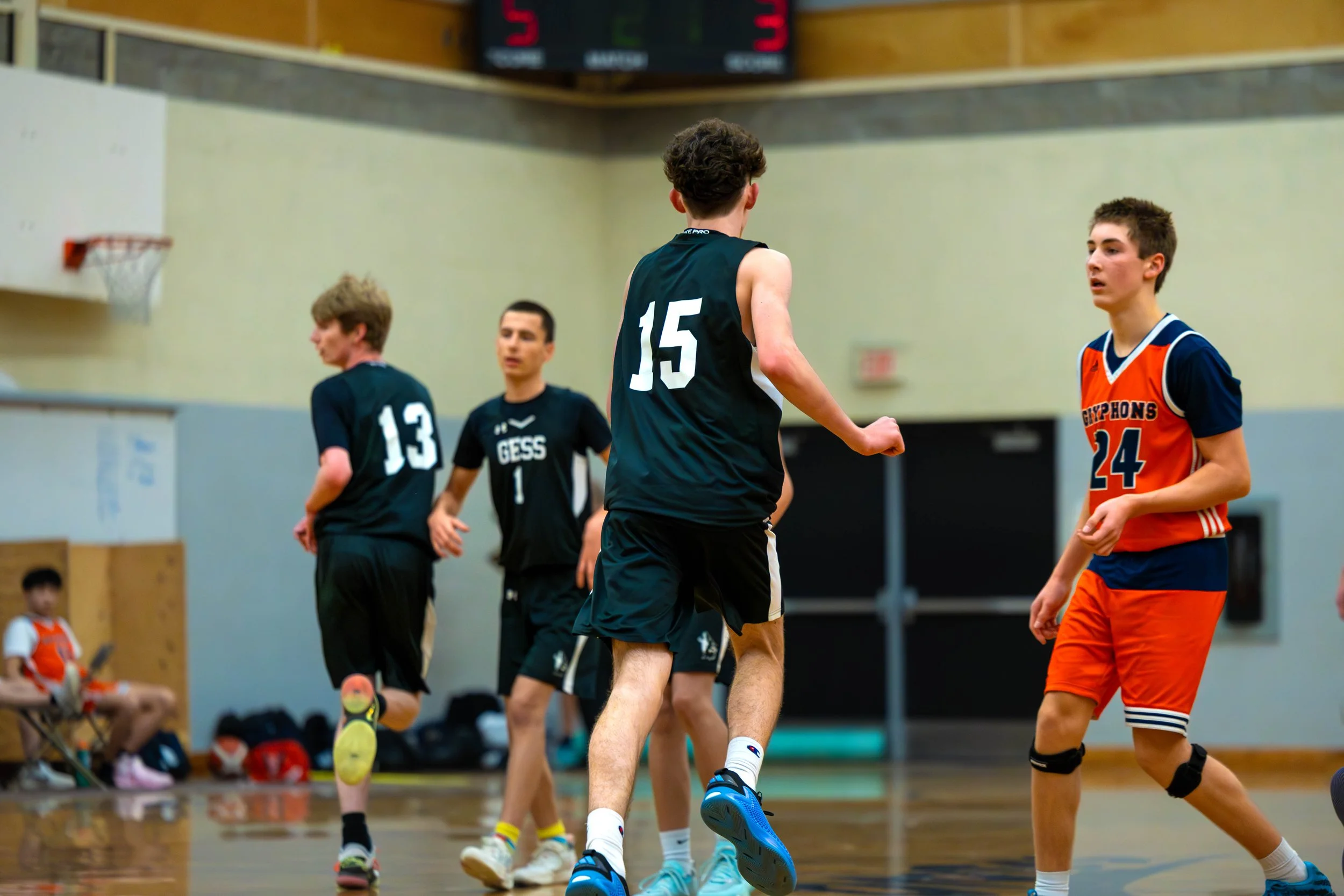 Young boys playing basketball indoors, with players wearing black and orange jerseys, one of whom is about to shoot or pass the ball, other players are positioned around the court.
