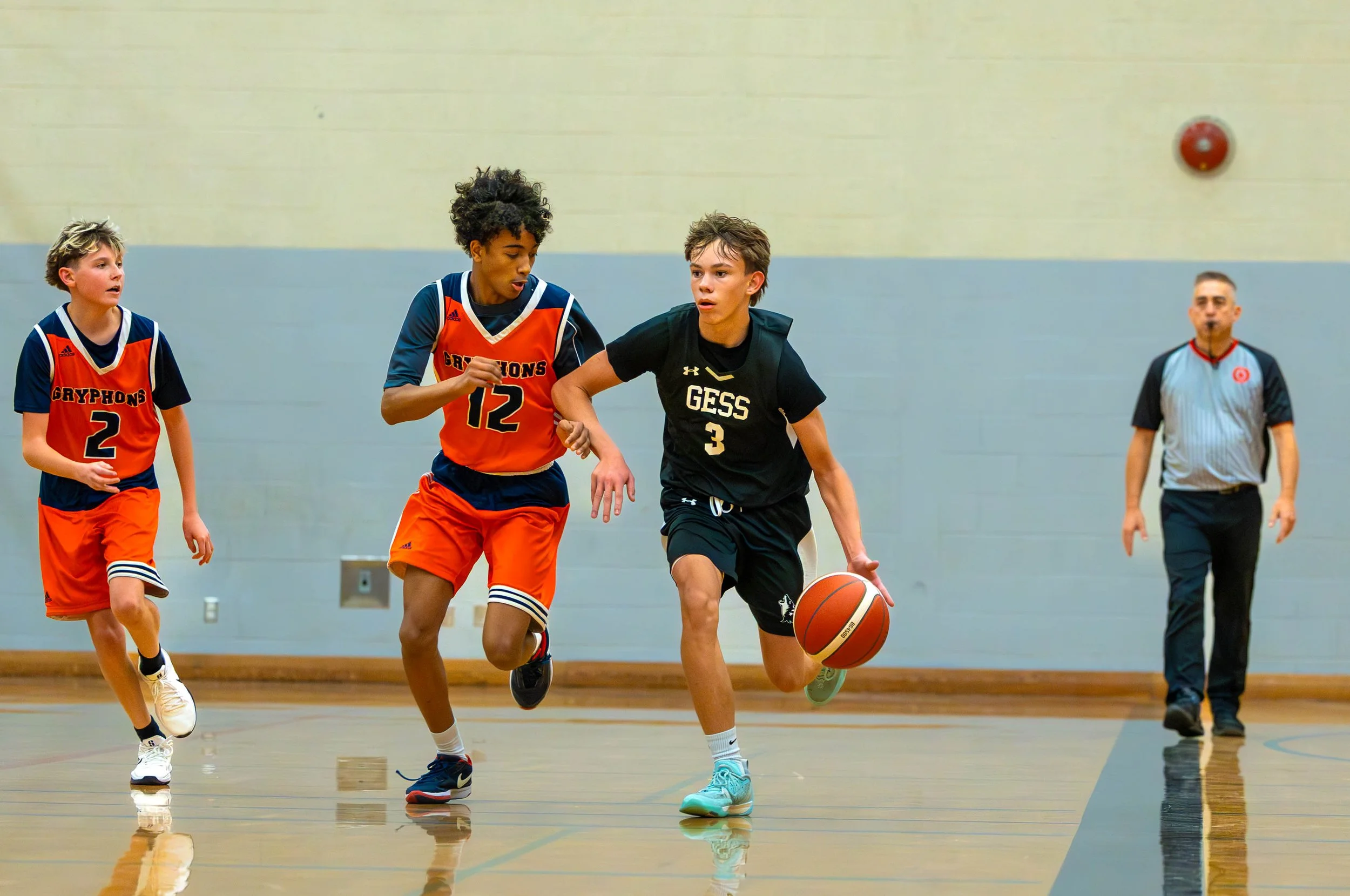Young boys playing basketball indoors, with one boy dribbling smoothly past two defenders, and a referee observing in the background.