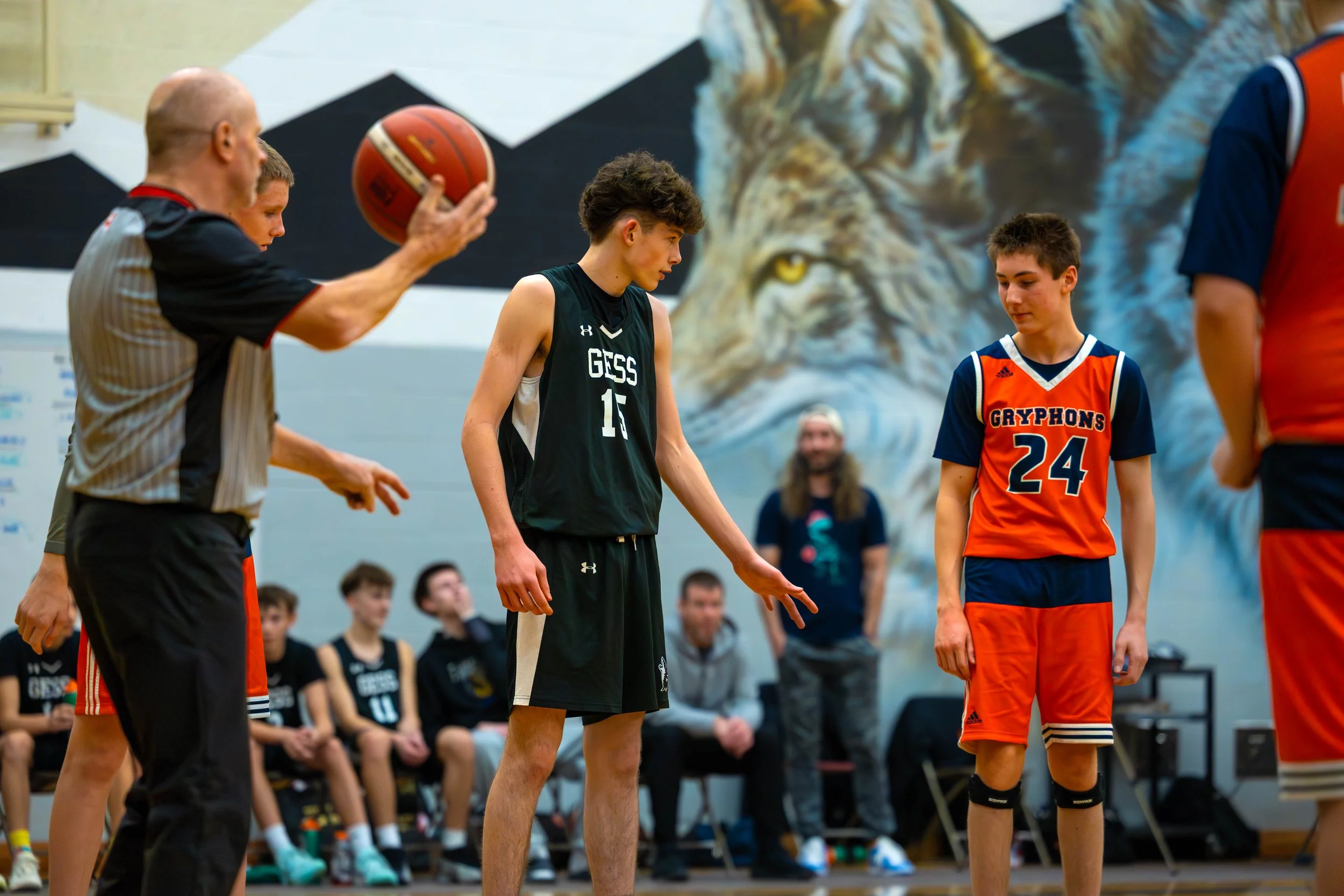 Young basketball players stand on court with referee holding a basketball, preparing for a game, with team benches and a mural of a lion in the background.
