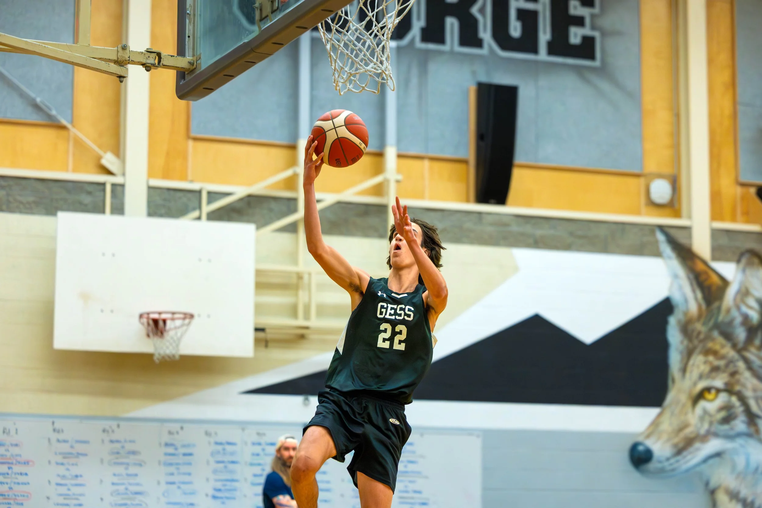 A young male basketball player in a black GESS jersey with the number 22 is leaping towards the basketball hoop, holding a basketball with both hands, preparing to make a shot in an indoor gymnasium.