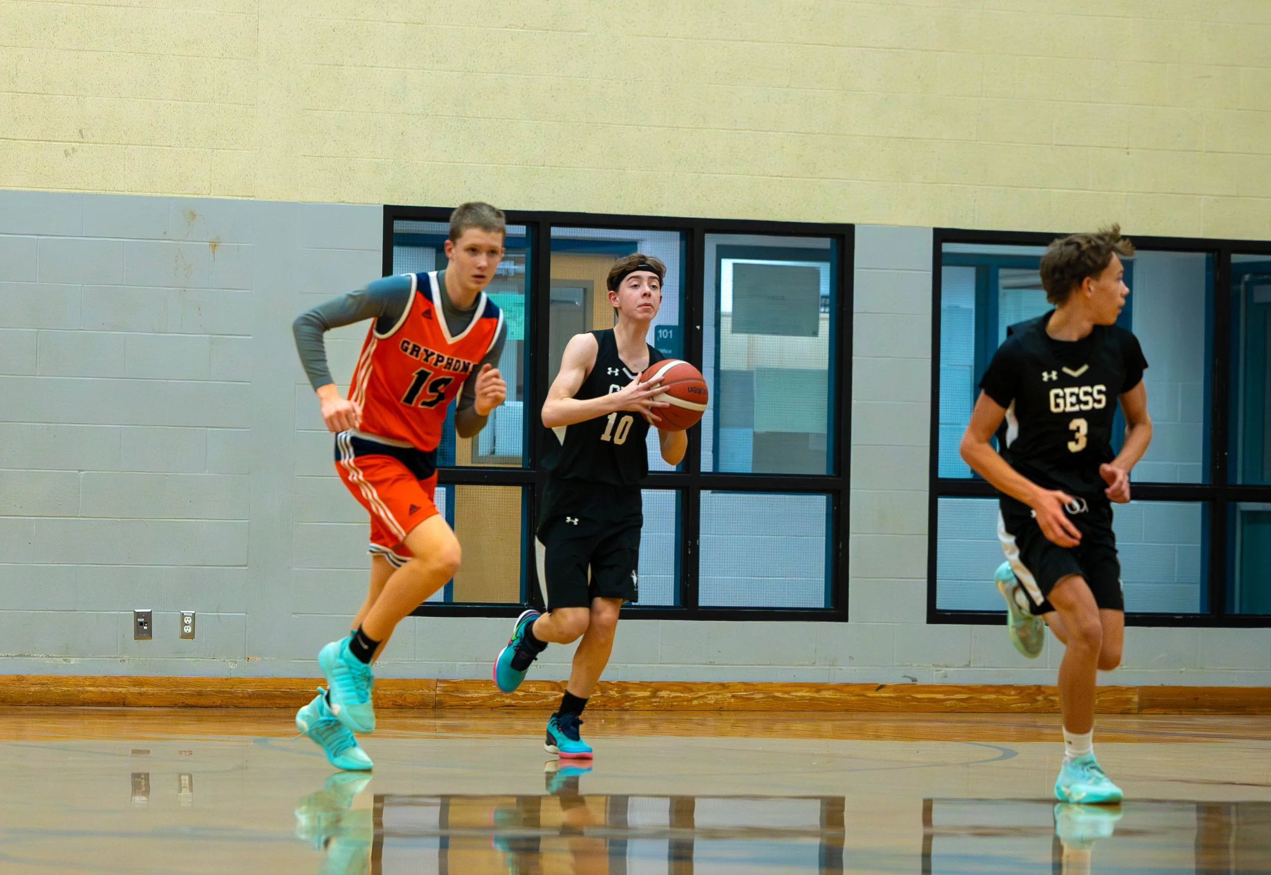 Three young male basketball players in a gym, with one holding a basketball, running and looking to the side.