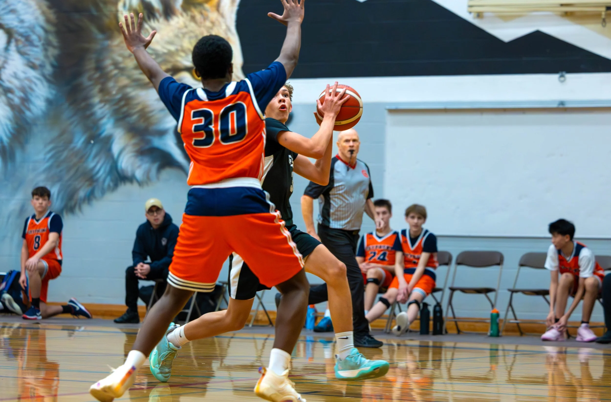 A youth basketball game with players on the court and bench, including a player in black holding the ball and a player in orange defending, with fans and coaches watching.