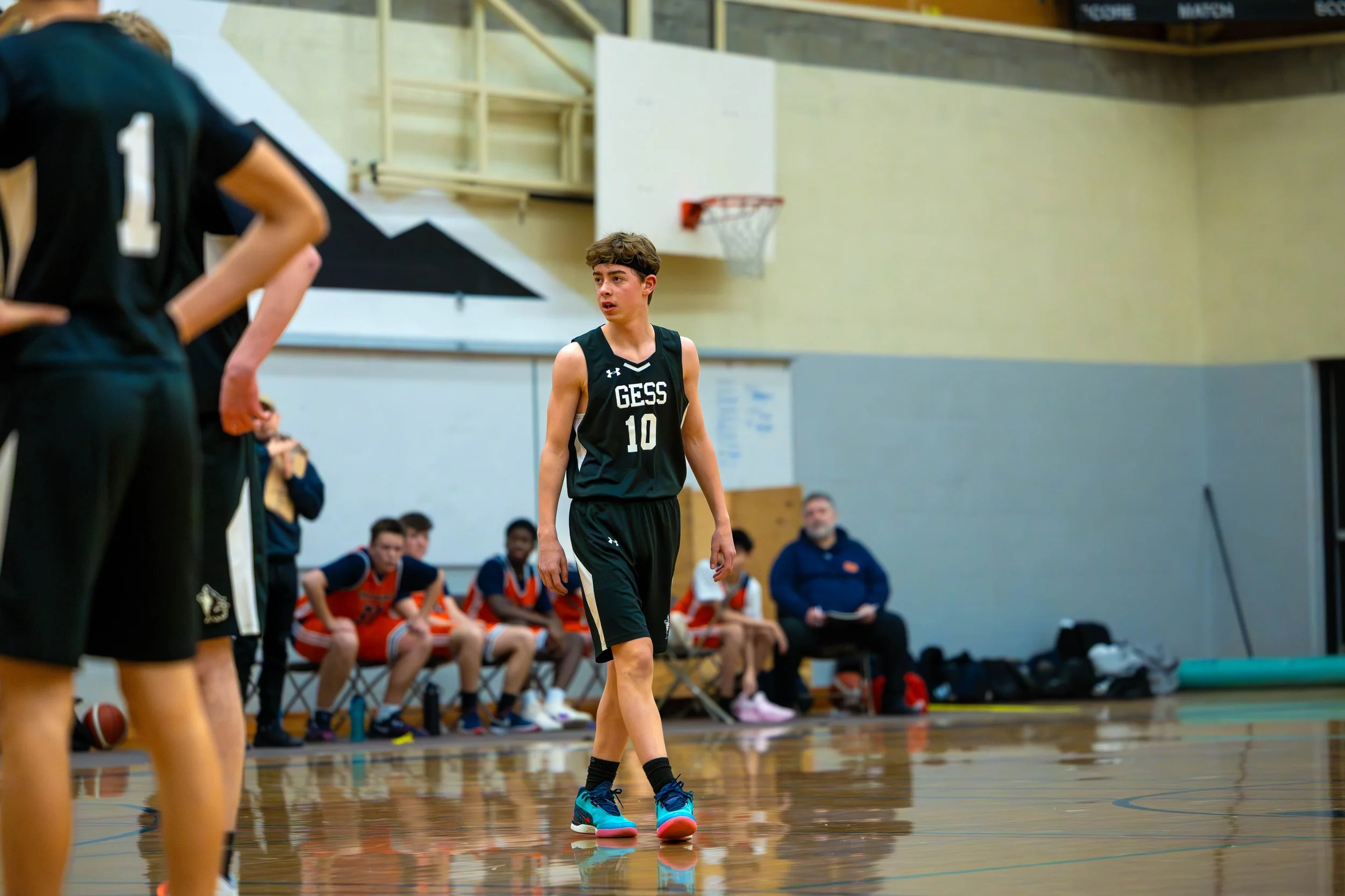 A young male basketball player wearing a black jersey with the number 10 and the word "GESS" stands on a basketball court during a game or practice, with teammates and a coach visible in the background.