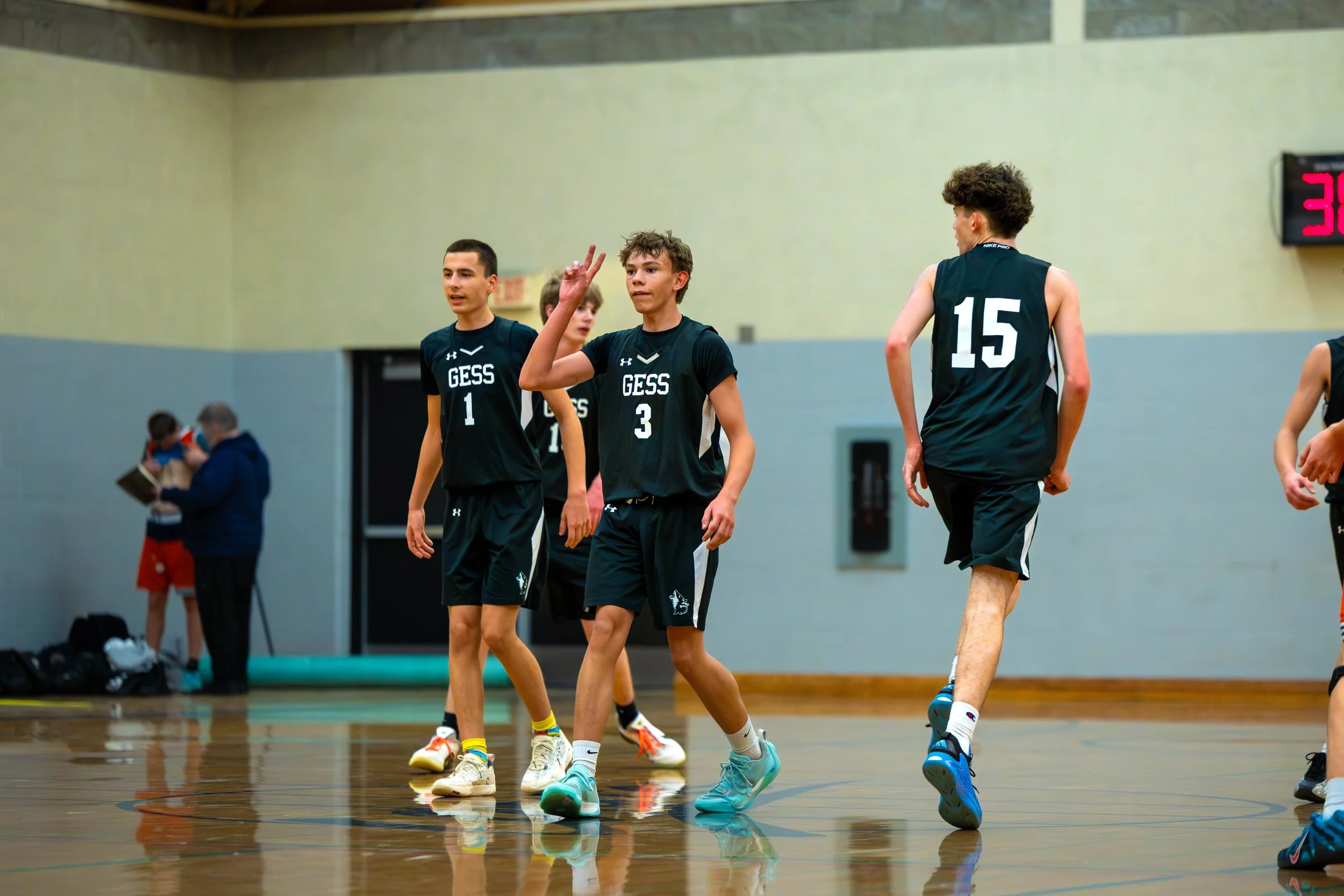 Youth basketball players on the court with one making a peace sign, wearing black uniforms with 'GESS' and numbers, in a gymnasium.