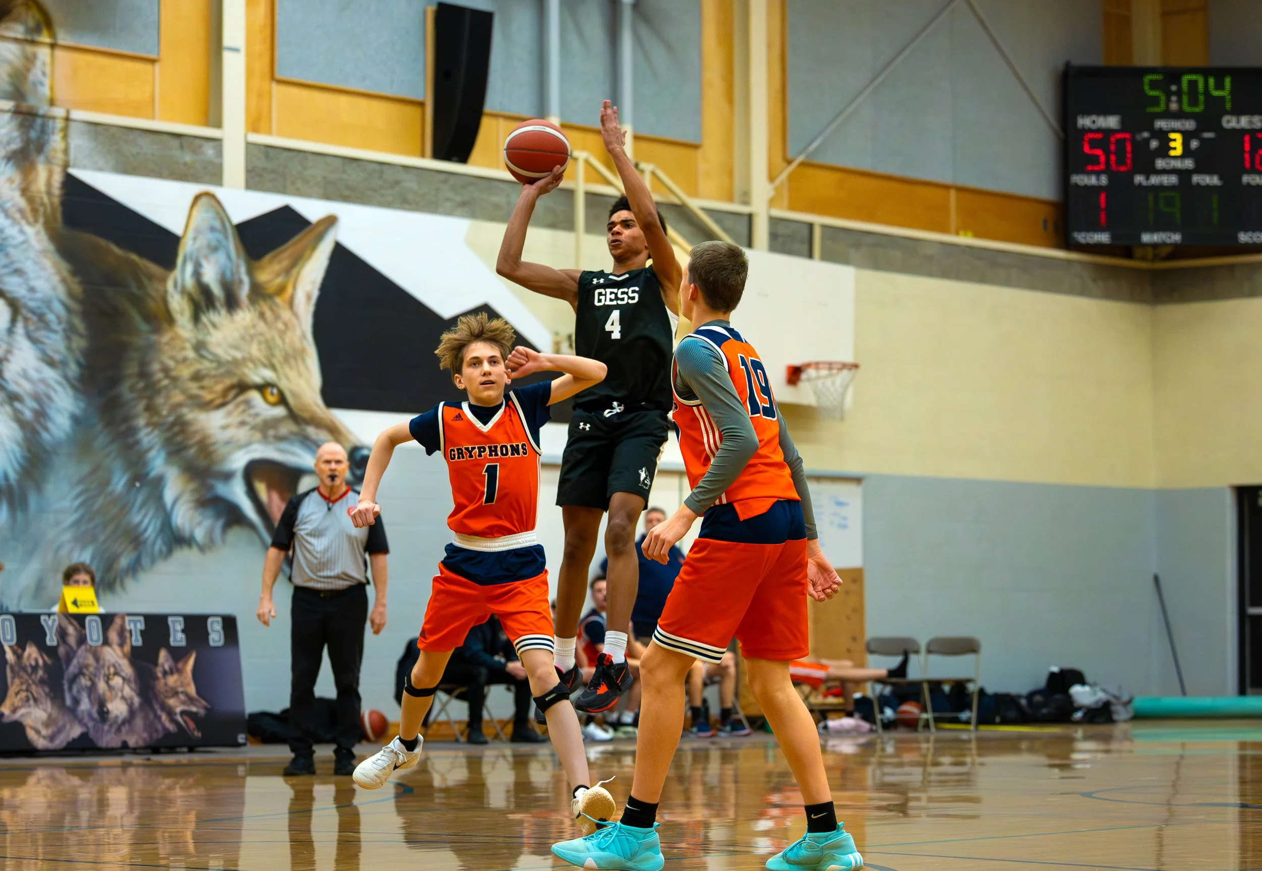 A boys' basketball game in progress with a player in black jumping to shoot the ball while two players in orange jerseys defend. The gym features a large wolf mural on the back wall and a scoreboard showing 5:04 remaining.
