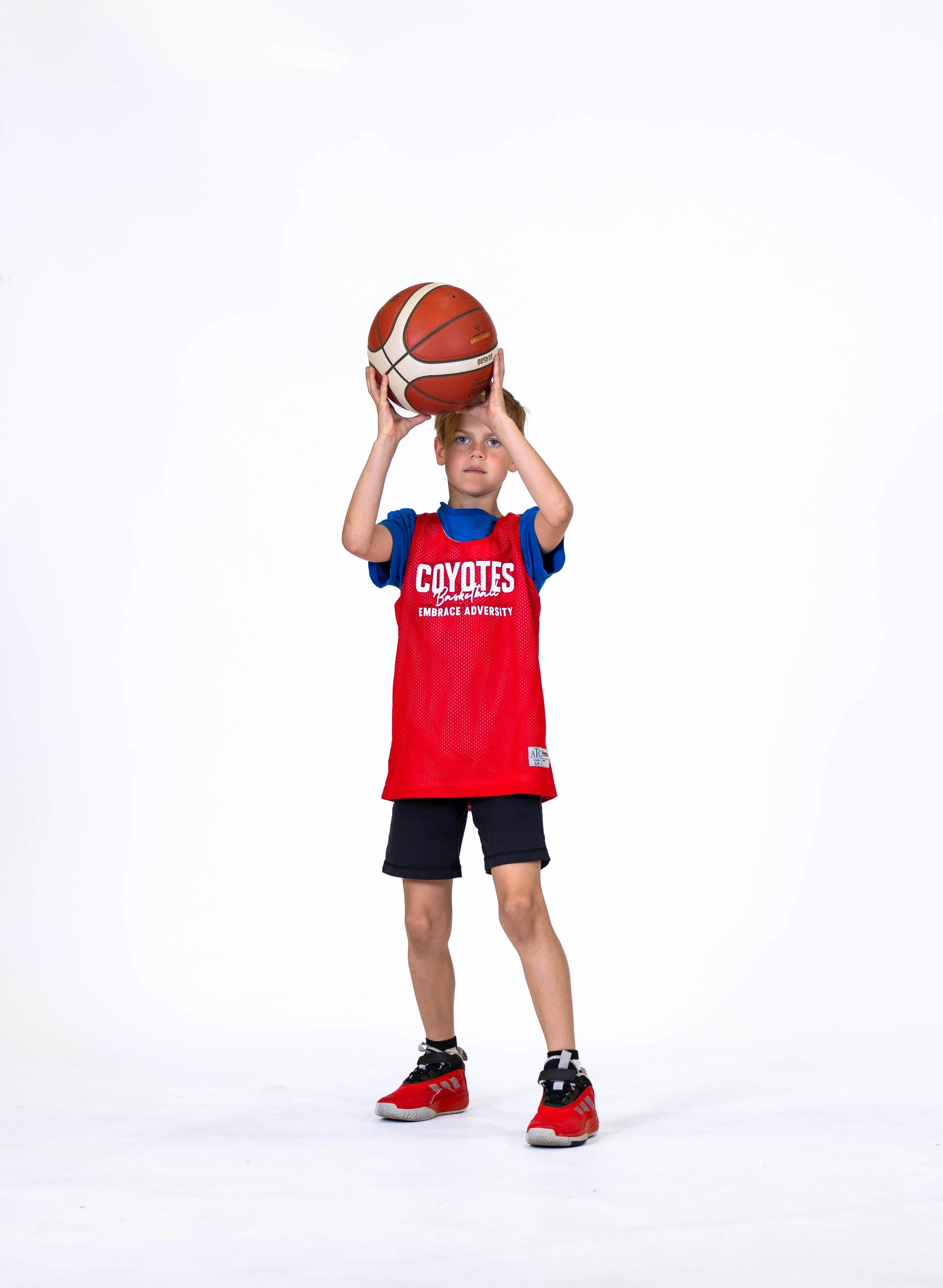 Young boy in a red basketball jersey with blue sleeves, holding a basketball above his head, standing in a studio with a white background, wearing red and black sneakers.