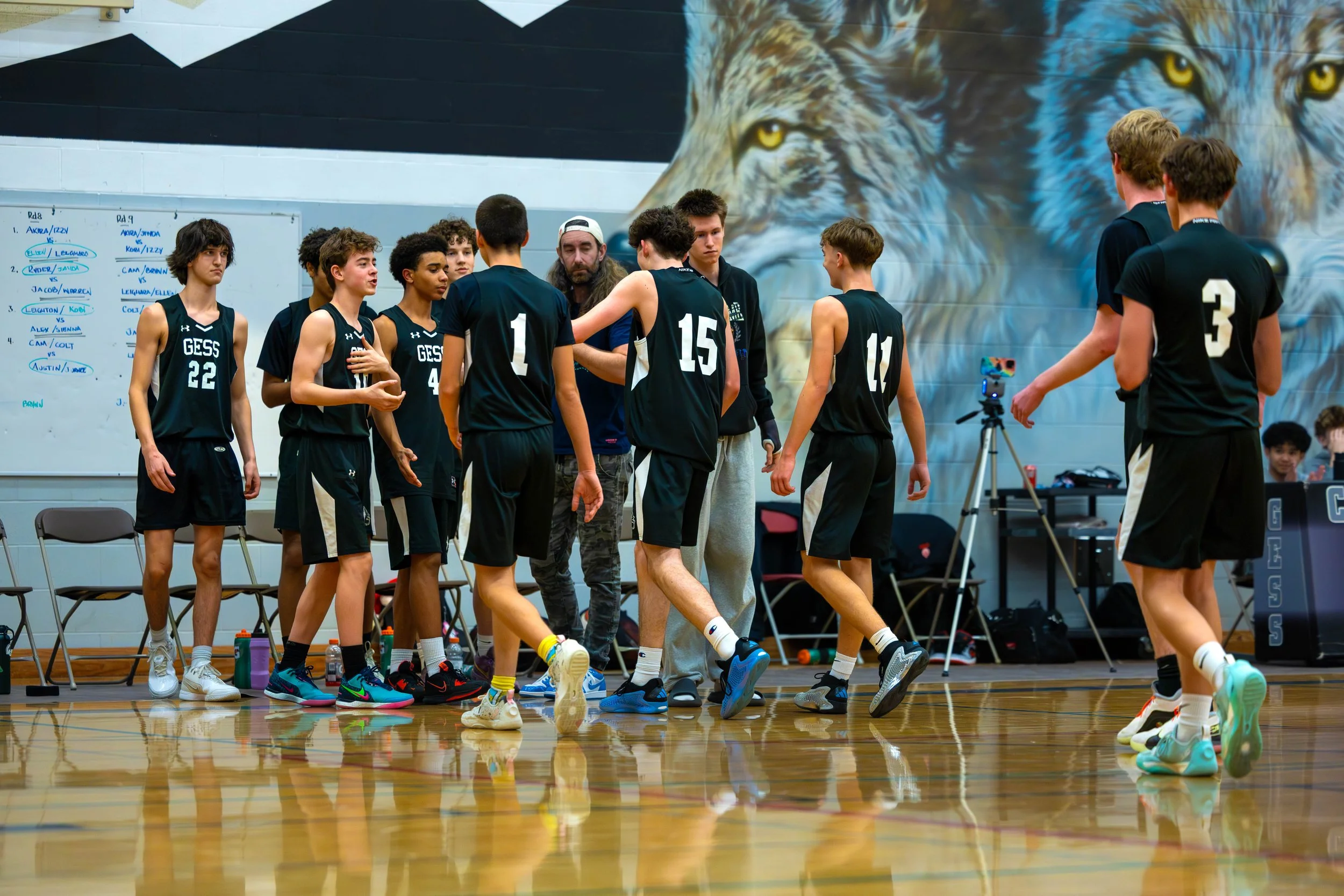 Basketball team in black jerseys and shorts gather on the court during a timeout or break, with a coach giving instructions, in a gym with a mural of a wolf on the wall.