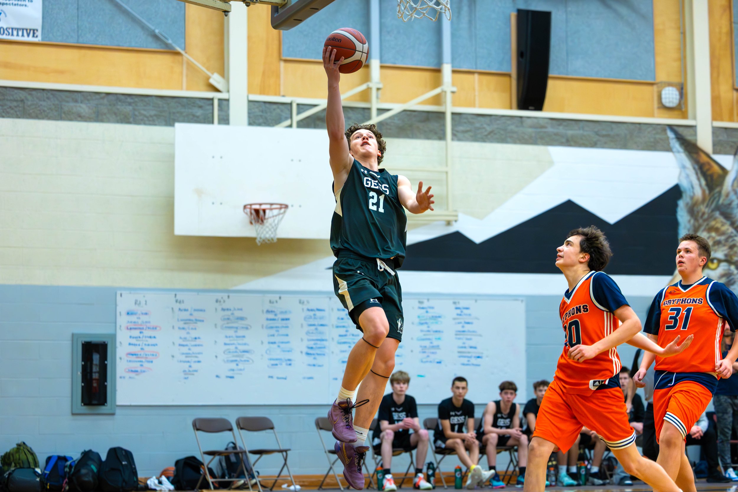A young basketball player in a black uniform jumps to shoot the ball in an indoor gymnasium, with two other players in orange uniforms nearby and a bench with seated players in the background.