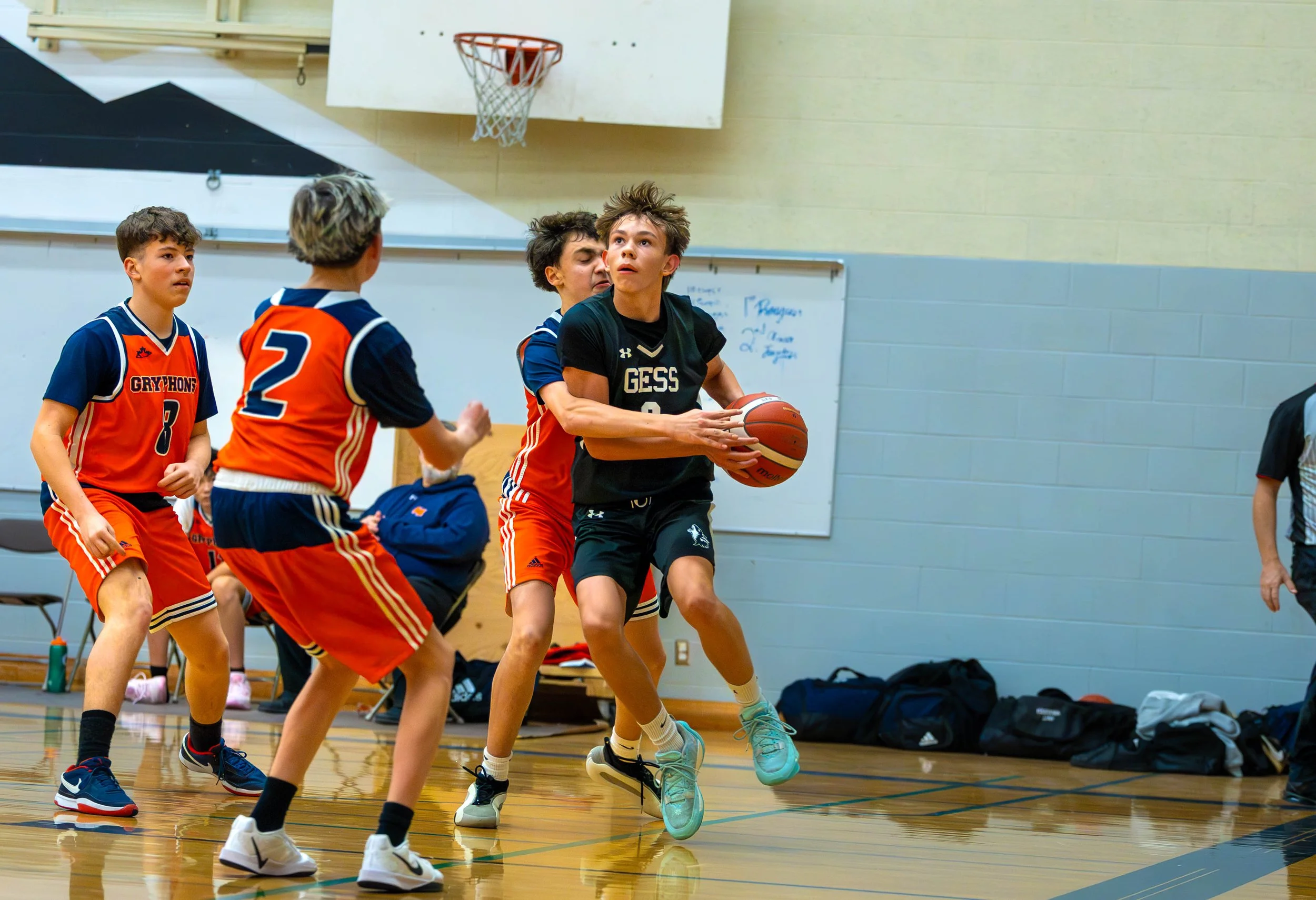 Young boys playing basketball in a gym. One boy in black is holding the ball, surrounded by three boys in orange jerseys.