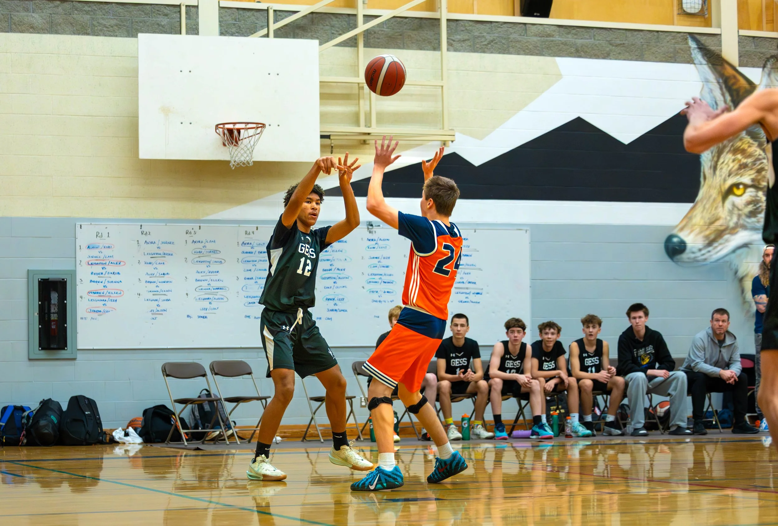 Two boys playing basketball inside a gym, one in black uniform shooting and the other in orange attempting to block, with seated players and coaching staff in the background.