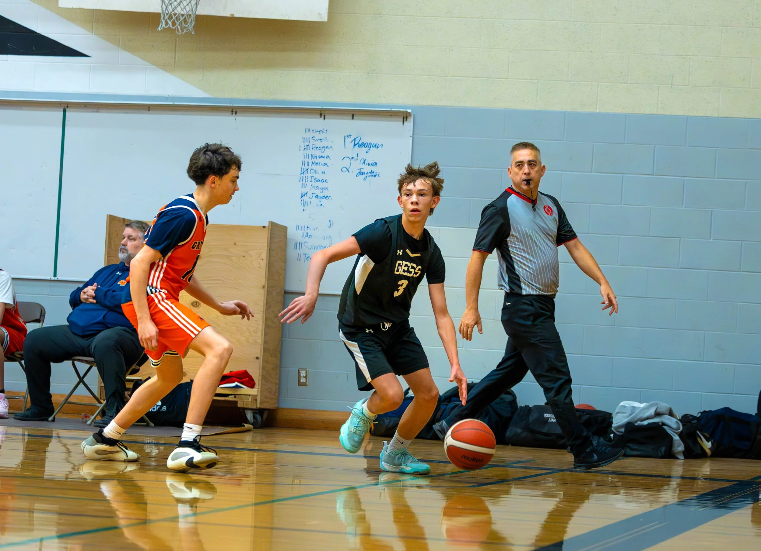 Young boy in black sports uniform dribbling a basketball on an indoor court while two other players and referee watch. One player is in an orange uniform and the referee is in a gray shirt with black pants.
