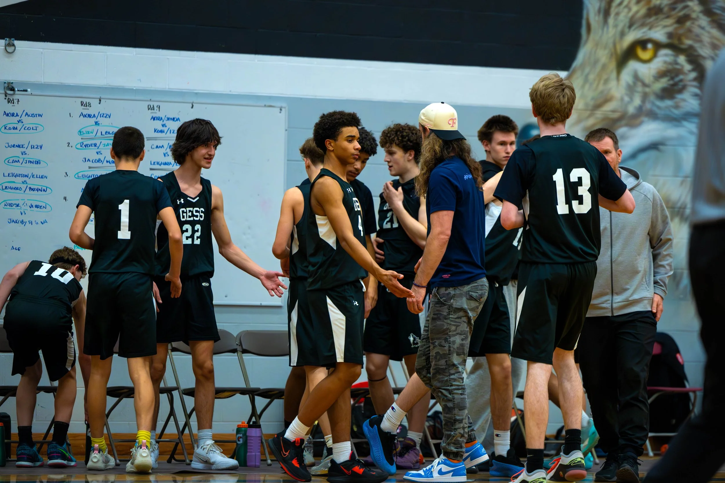 A group of young male basketball players in black jerseys shake hands with a coach during a game break in a gymnasium.