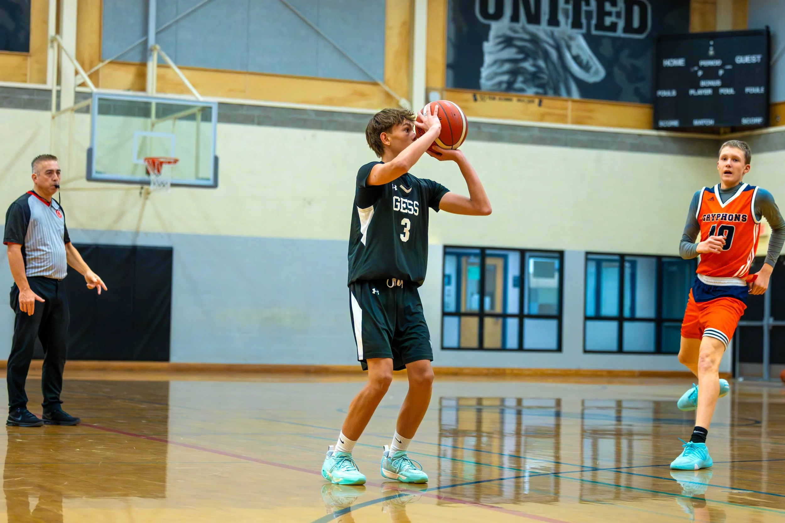 Two young boys playing basketball indoors with a referee in the background, one preparing to shoot the ball, the other running.