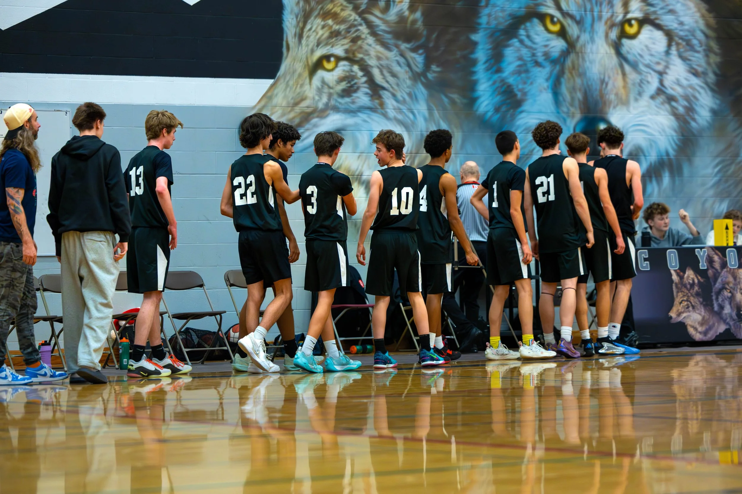 A high school basketball team in black jerseys standing in a gymnasium, with a large mural of wolves on the wall behind them.