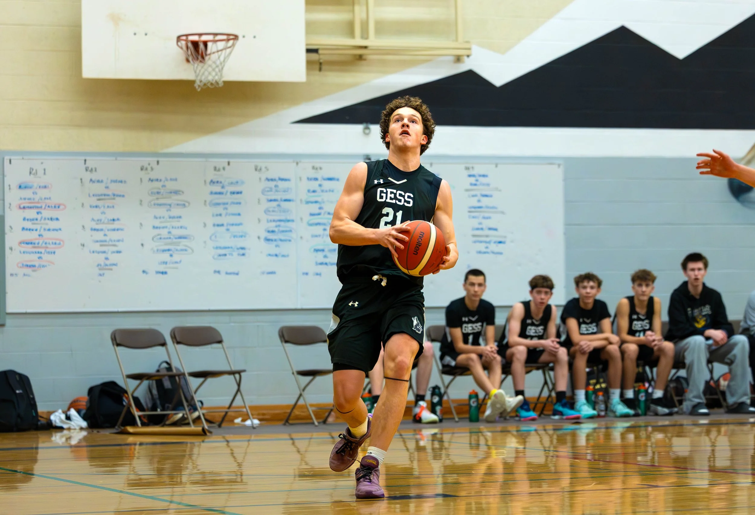 A young man in a black basketball uniform with the number 21 is playing basketball on an indoor court, holding a basketball in both hands, with a group of players sitting on the bench behind him and a whiteboard with writing on the wall.