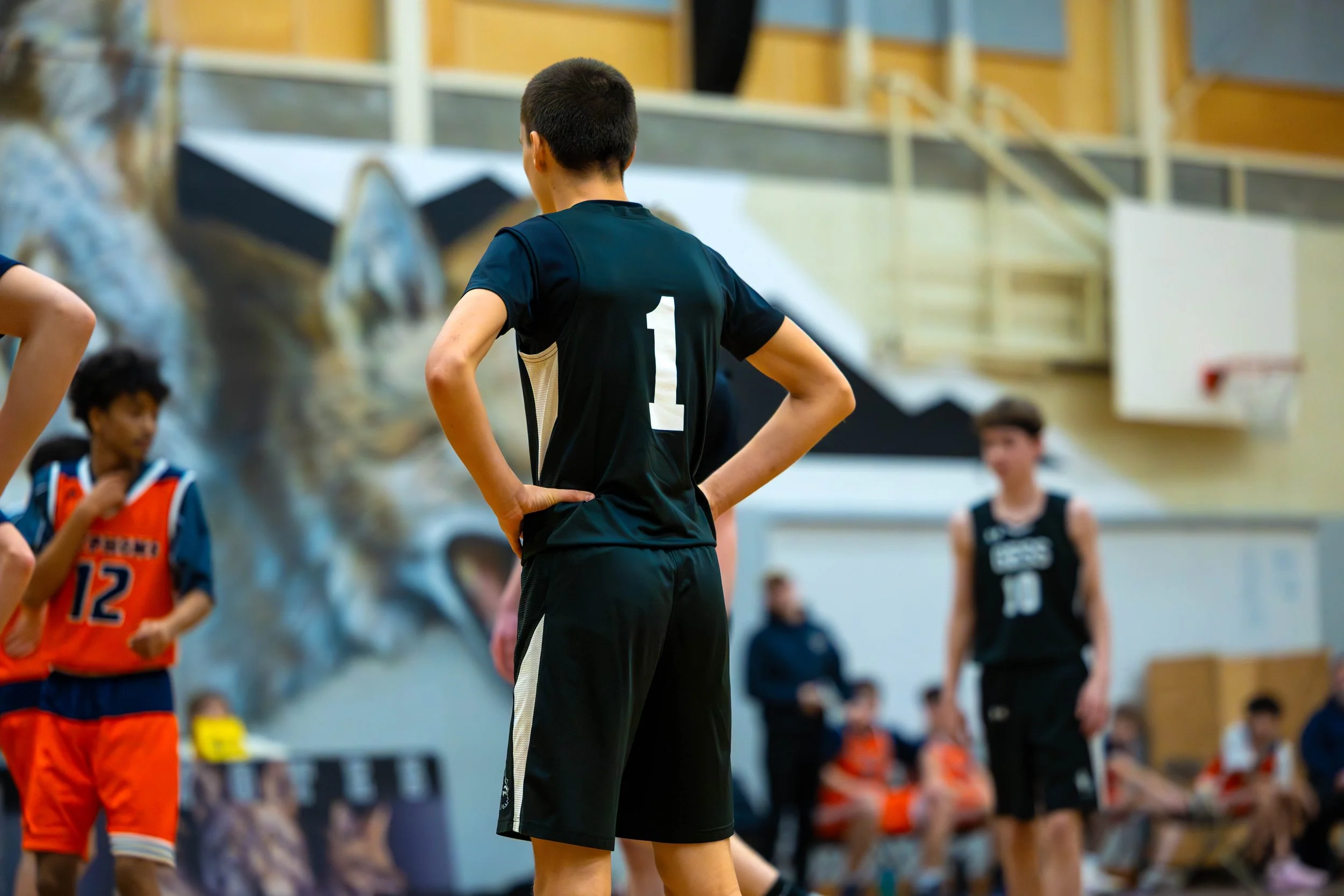 A group of young basketball players on a court, with one player in black jersey numbered 1 standing with hands on hips, others in the background, and a large mural of a wolf painted on the wall.