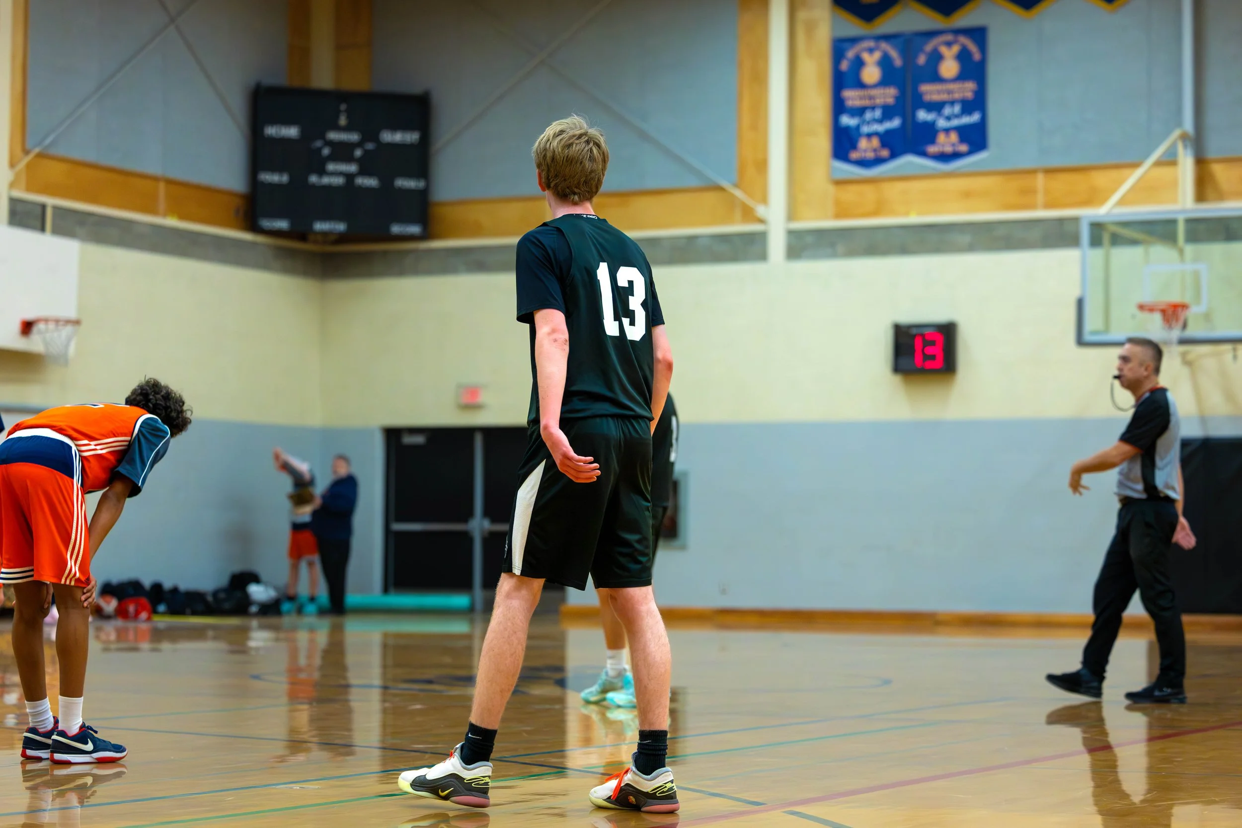 A basketball game in progress at indoor gym with players and referee, and a digital score showing 13 points.
