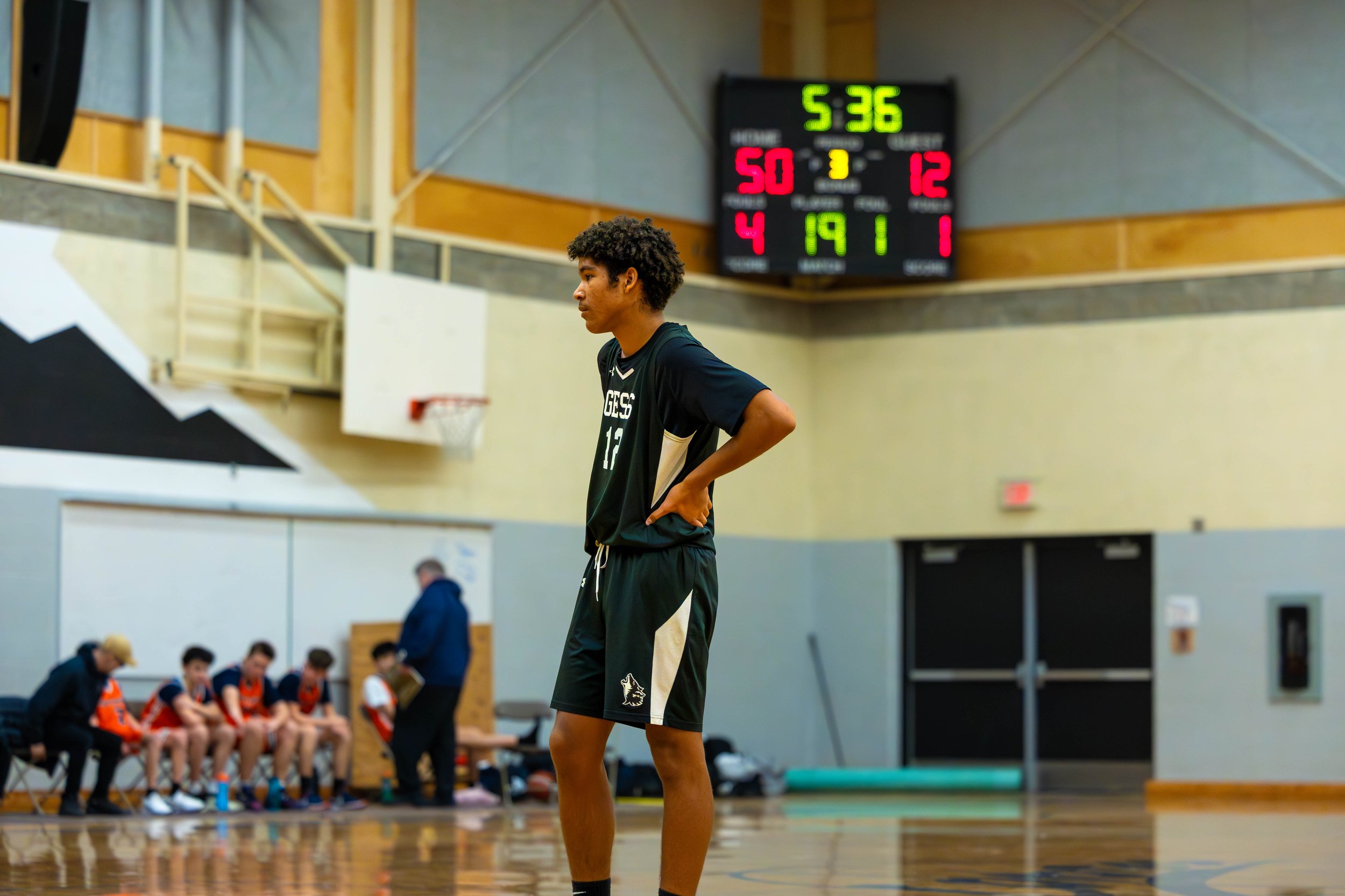 A young male basketball player standing on the court during a game, with his hands on his hips. He is wearing a dark sports uniform, and there are other players and a coach sitting on the bench in the background. The scoreboard shows 5 minutes and 36