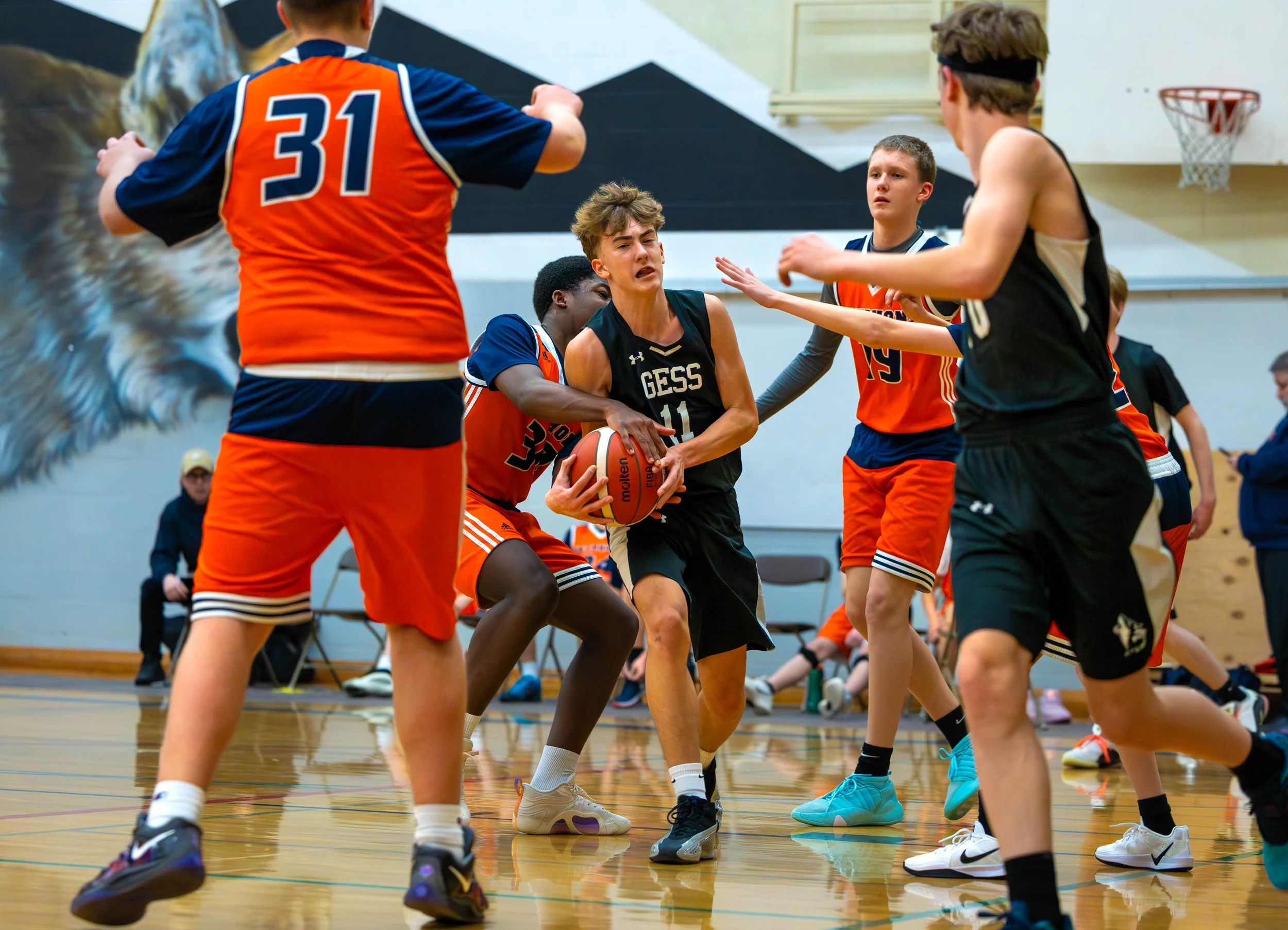 Young boys playing basketball in an indoor court. One boy in a black jersey is holding the basketball and is being guarded by other players. The scene shows intense action during the game.