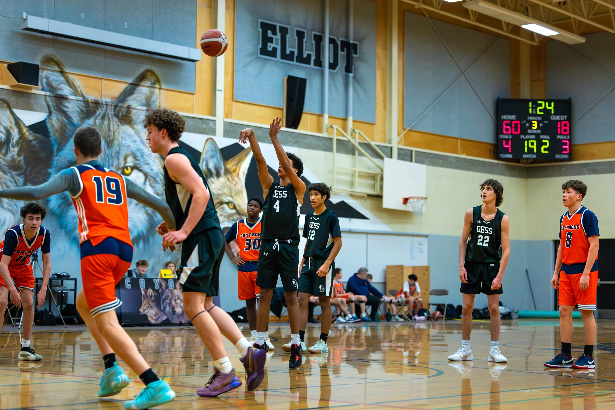 Young boys playing basketball on an indoor court with a large mural of a wolf on the wall and an electronic scoreboard showing a score of 4 to 3 with 1 minute and 24 seconds remaining.