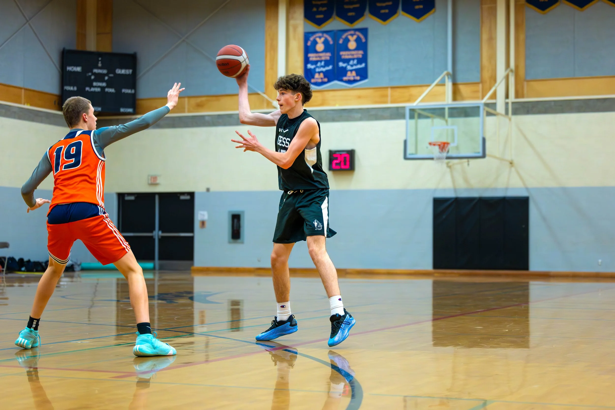 Two boys playing basketball in an indoor gym, one in a black jersey shooting the ball, the other in an orange jersey defending.