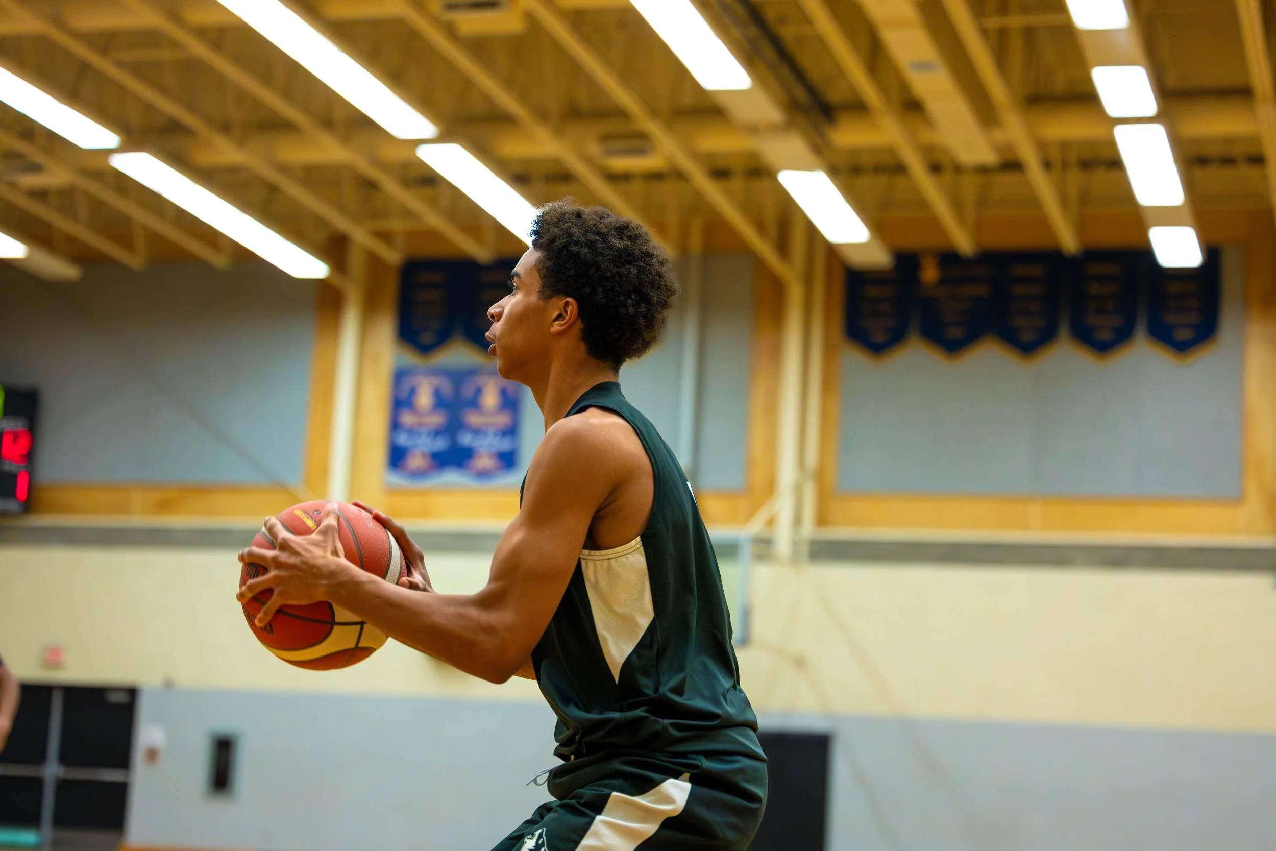 A young man in a dark sports jersey prepares to shoot a basketball on an indoor court with a wooden ceiling and blue banners.