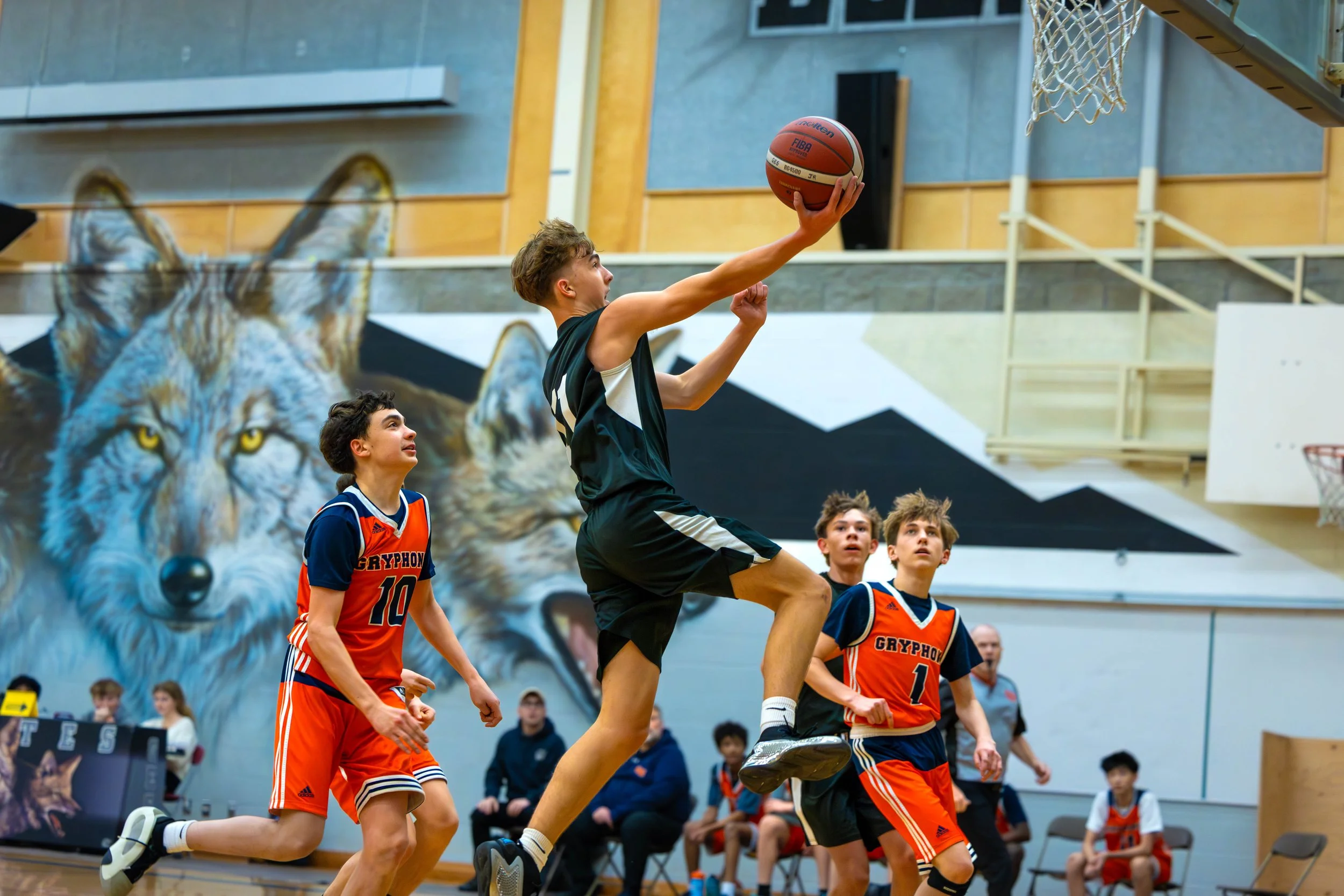 Young basketball players compete during a game; one player in black jumps to score while others in blue and orange look on.