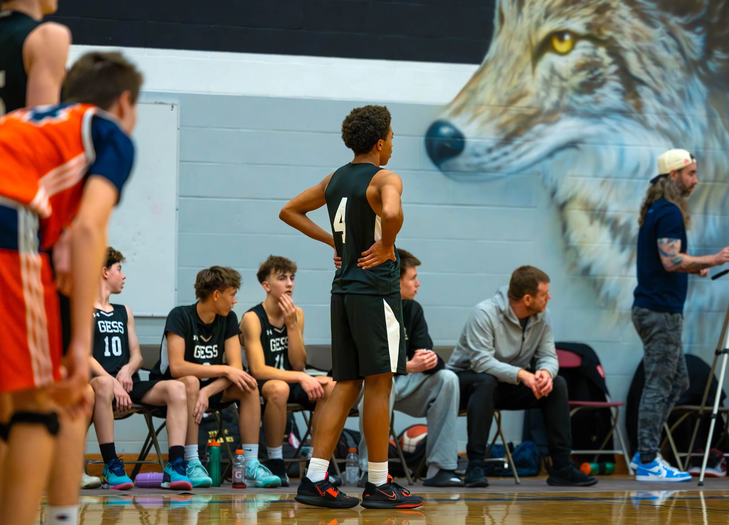 A basketball team sitting on the bench during a game, with players in black jerseys marked 'GESS', while a player in a black jersey with the number 4 stands with hands on hips, watching the game on the court. A coach and a man with long hair and a ca