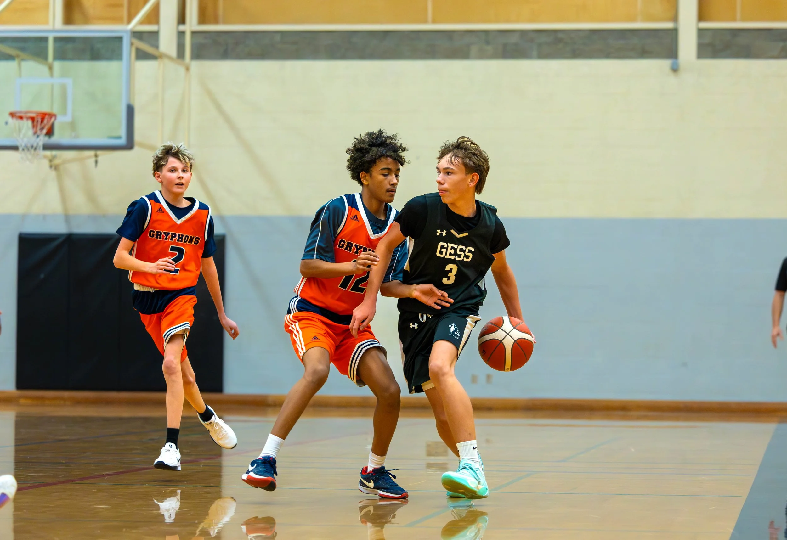 Three boys playing basketball in an indoor gym. One boy in a black and gold uniform dribbling the ball while two others in orange and blue uniforms try to block him.
