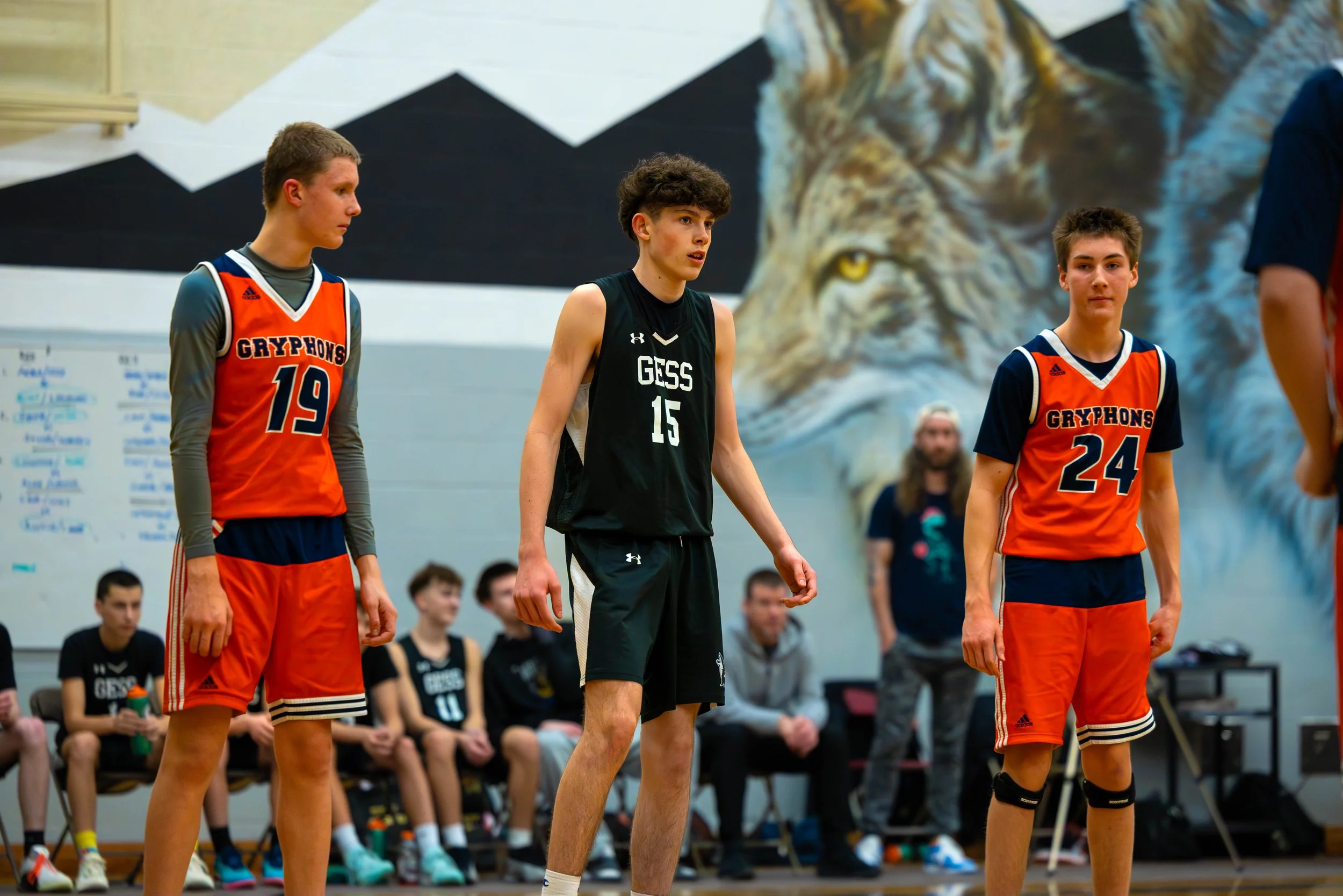 Three basketball players standing on a court, with a team bench and a large lion mural in the background.