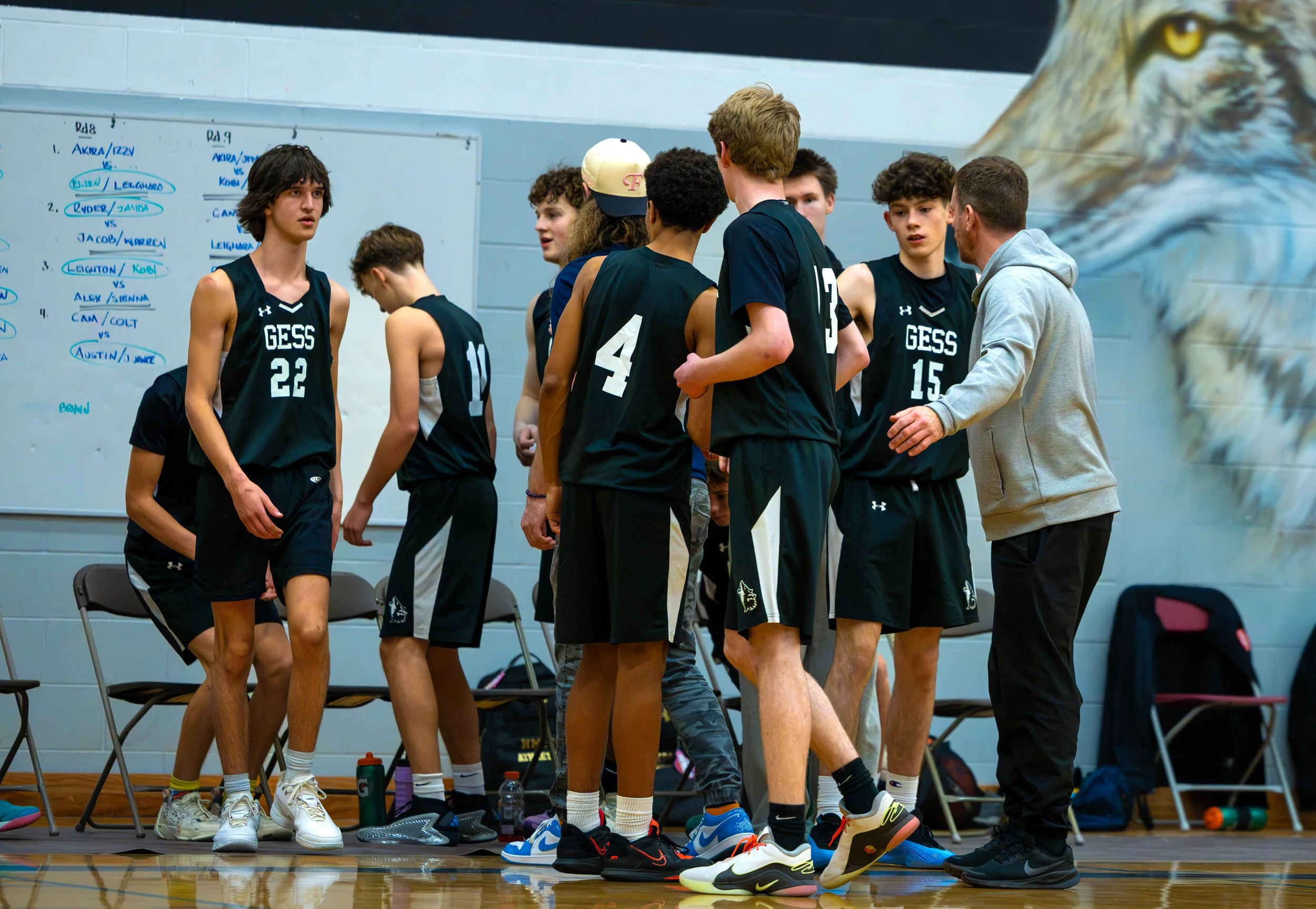 A group of young basketball players and their coach huddled on a basketball court during a game or practice. They are wearing black jerseys with 'GESS' written on them, and some are listening attentively to the coach. A large mural of a bird's head i
