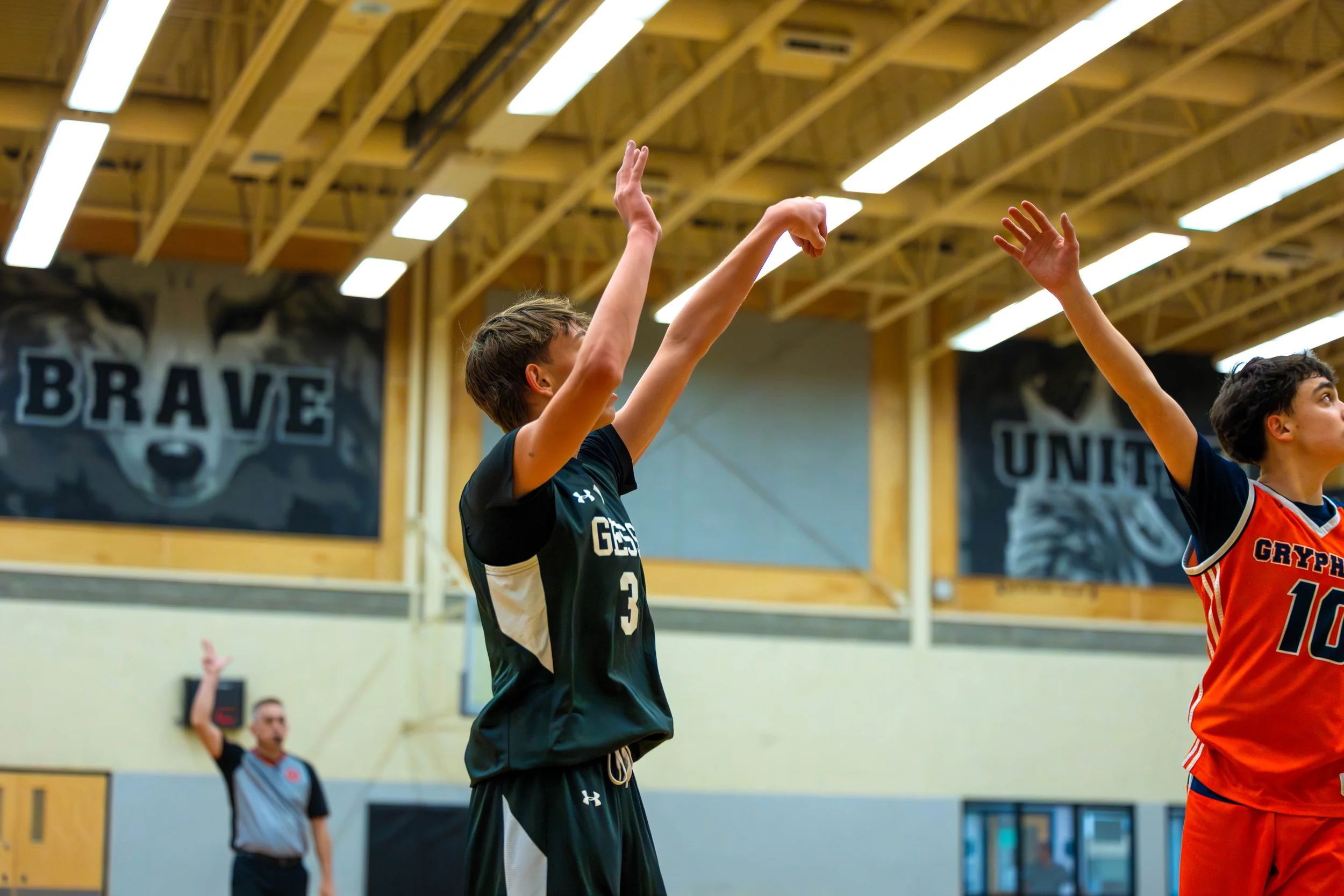 Two young male basketball players shooting free throws during a game in an indoor gym with wooden beams on the ceiling, with banners on the walls that say 'BRAVE' and 'UNITED'.