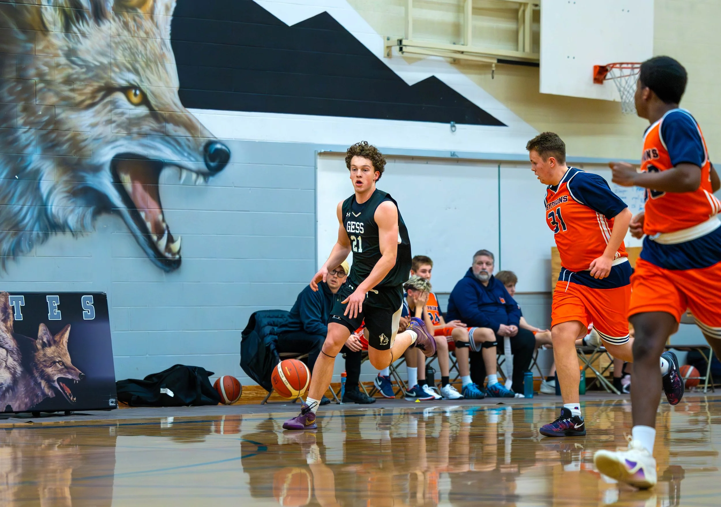 A young basketball player dribbling the ball during a game in an indoor gym, with teammates and coaches sitting on the bench in the background. The gym features a mural of a roaring wolf on the wall.