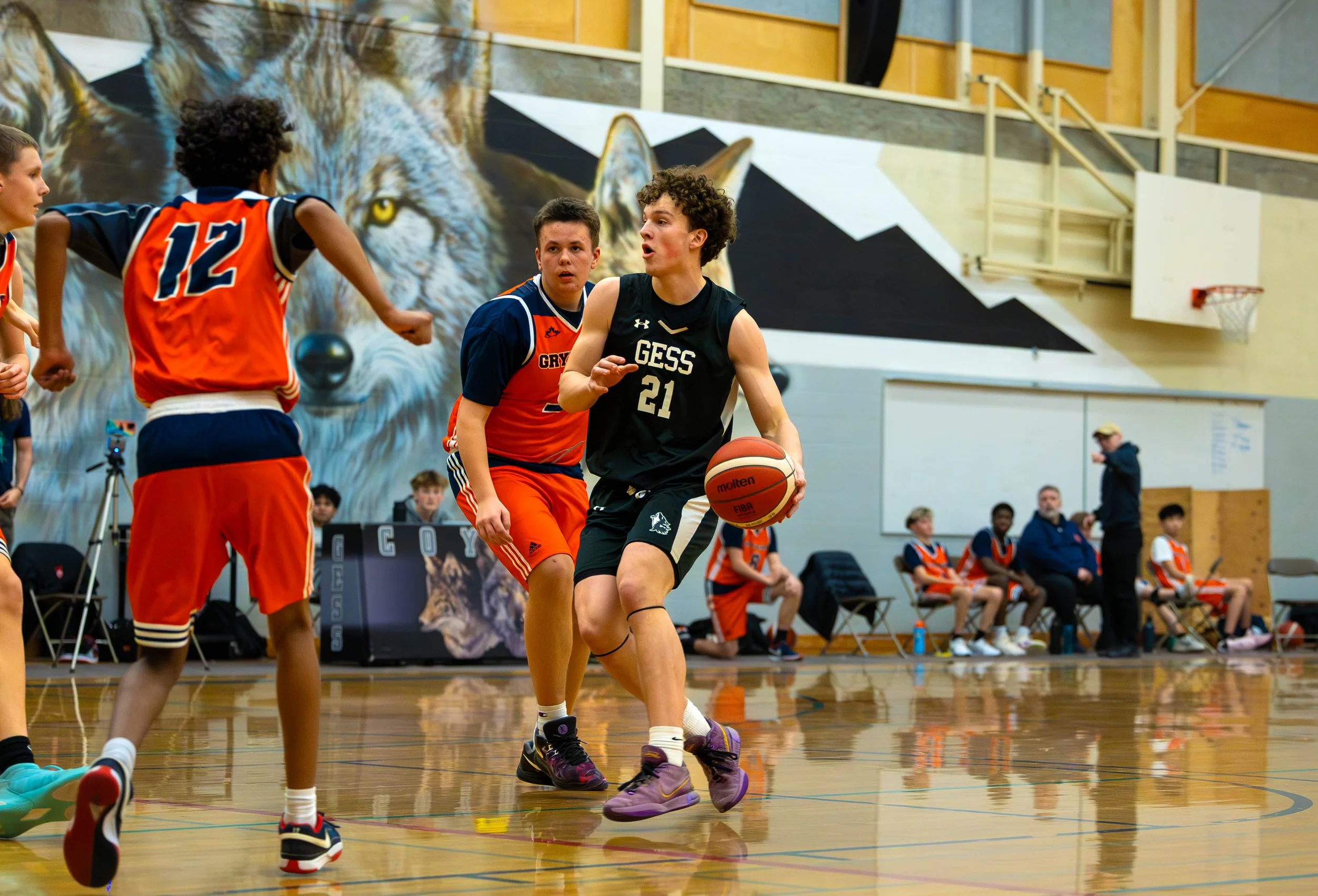 Young basketball players in uniform on court, with one dribbling the ball while others defend, in a gymnasium with a lion mural on the wall.