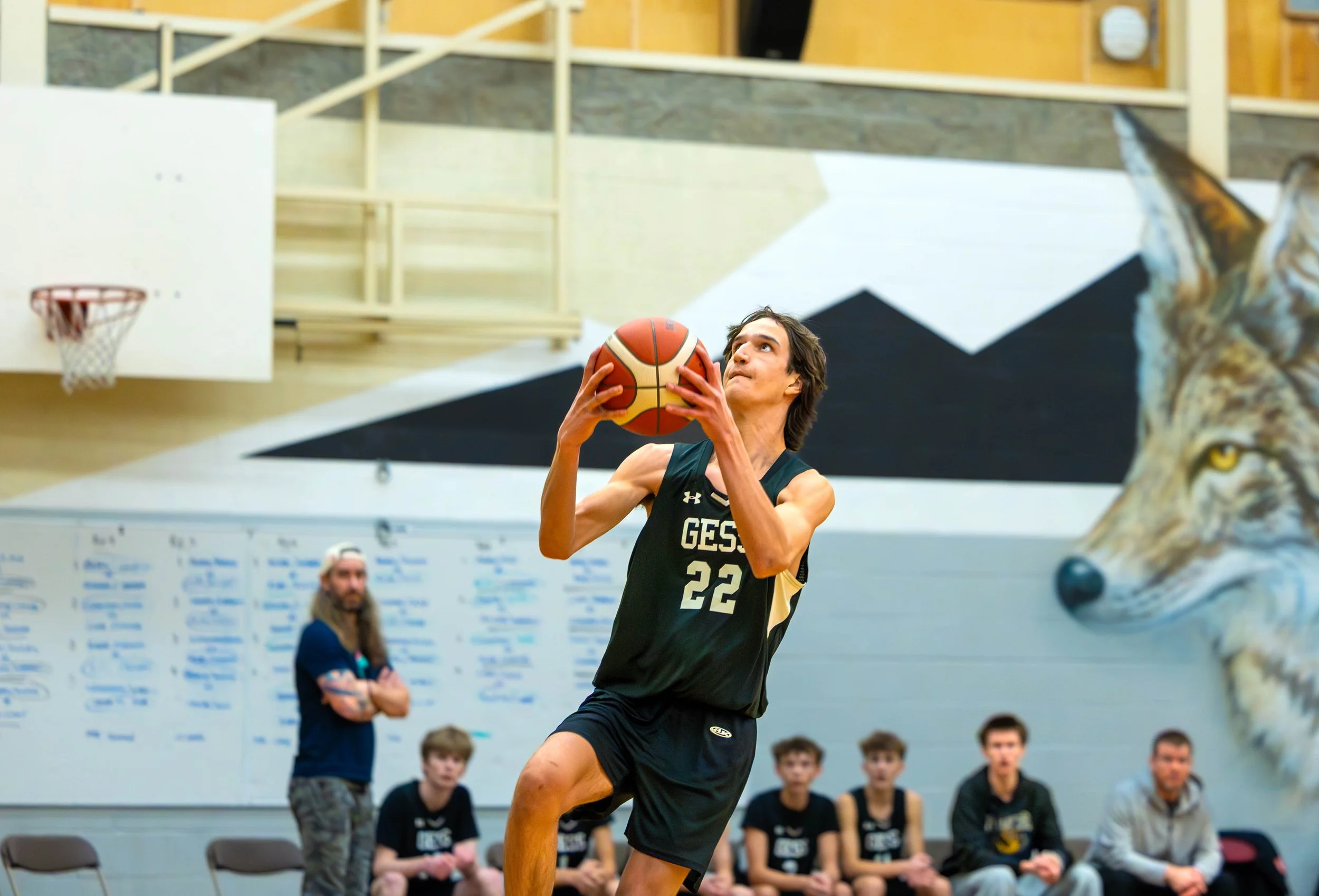 A basketball player in a black and gold uniform with number 22 is going for a shot in an indoor gym, with a wolf painted on the wall and several spectators sitting and standing nearby.