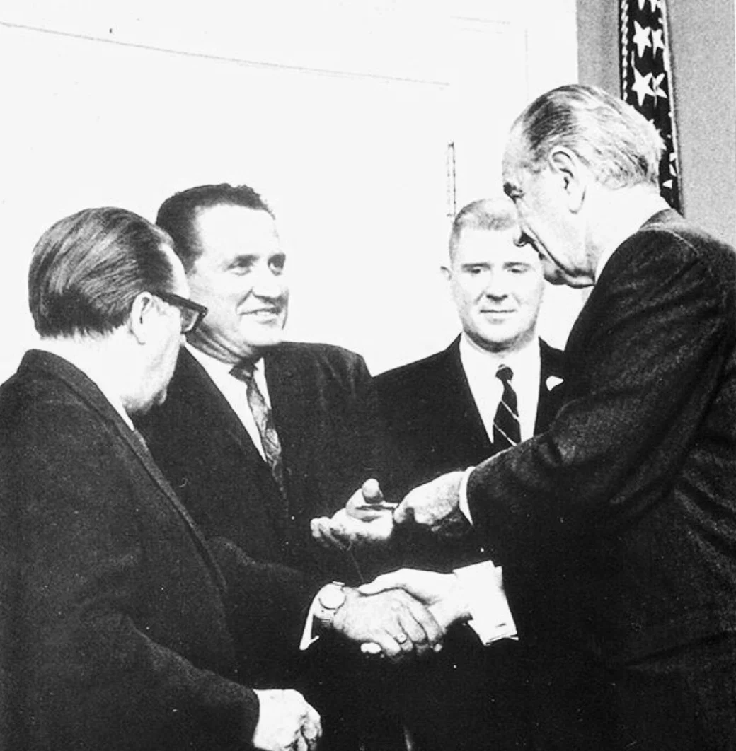 U.S. President Lyndon B. Johnson hands Washington State Insurance Commissioner Karl Herrmann the pen used to sign a pro-consumer law advocated by Herrmann. Washington Senator Warren Magnuson stands beside Herrmann on the left.