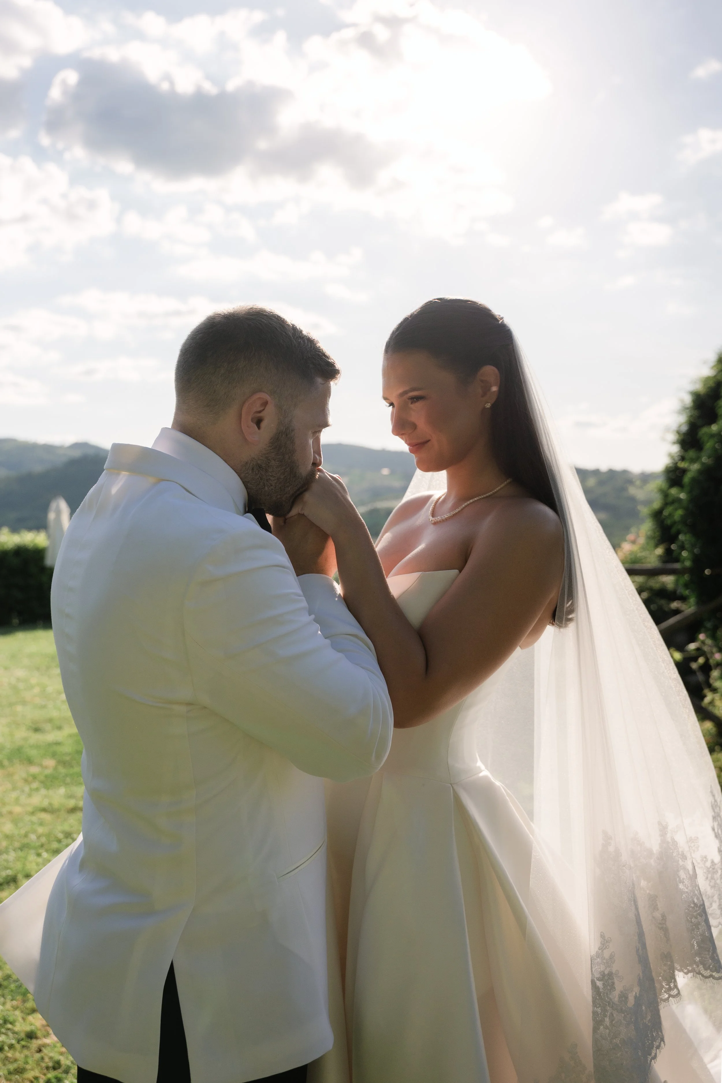 A couple dressed in wedding attire standing outdoors on a sunny day. The man in a white tuxedo is holding hands with the woman in a strapless wedding gown with a long veil. They are smiling and-looking at each other. The background shows a scenic landscape with green hills, trees, and a sky with clouds.