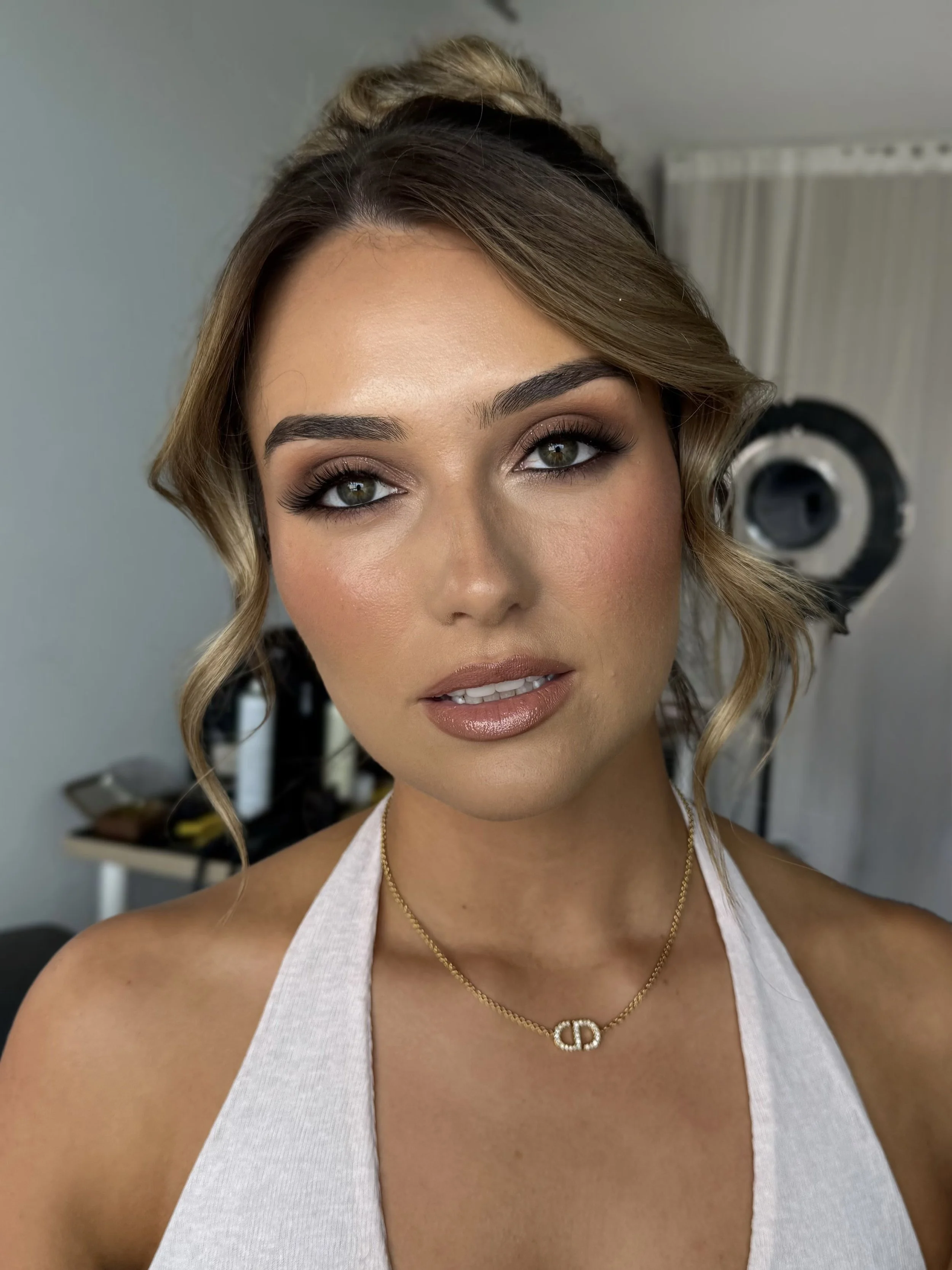 Close-up of a woman with styled hair, makeup, and a gold necklace with a 'CD' pendant, wearing a white halter top, standing in a room with a light green wall and a hairdryer in the background.