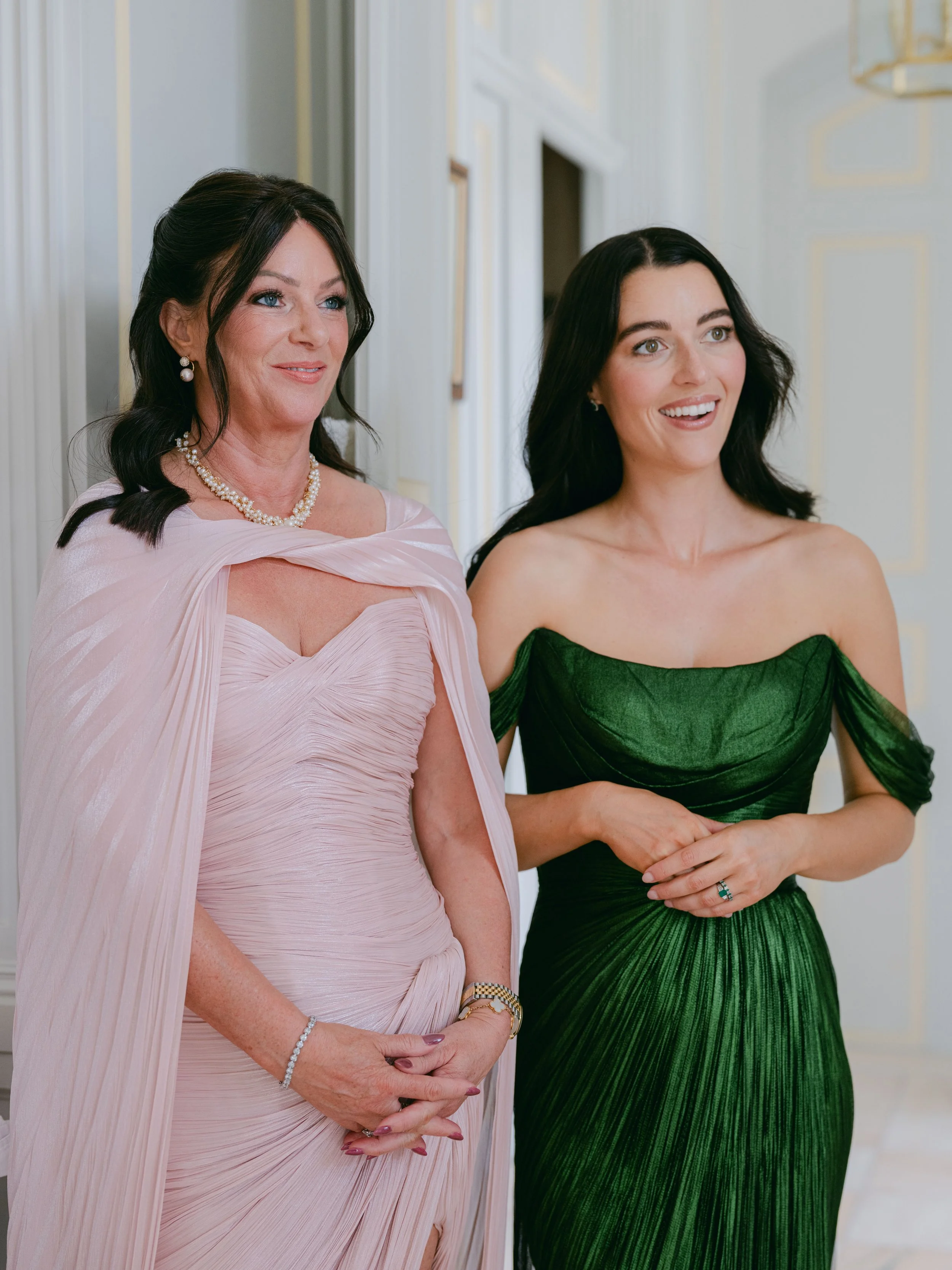 Two women at a formal event, one wearing a pink dress and the other in a green dress, smiling and standing close together indoors.
