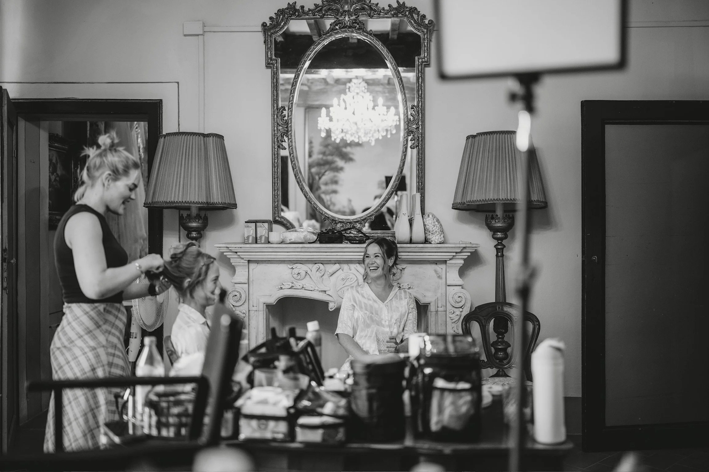 A woman is sitting on a chair in front of a fireplace, smiling. Two other women are standing nearby, one styling the woman's hair. There are lamps and vases on the fireplace mantle, and a chandelier is reflected in the mirror above it. The setting appears to be a salon or dressing room, with various items on a table in the foreground.
